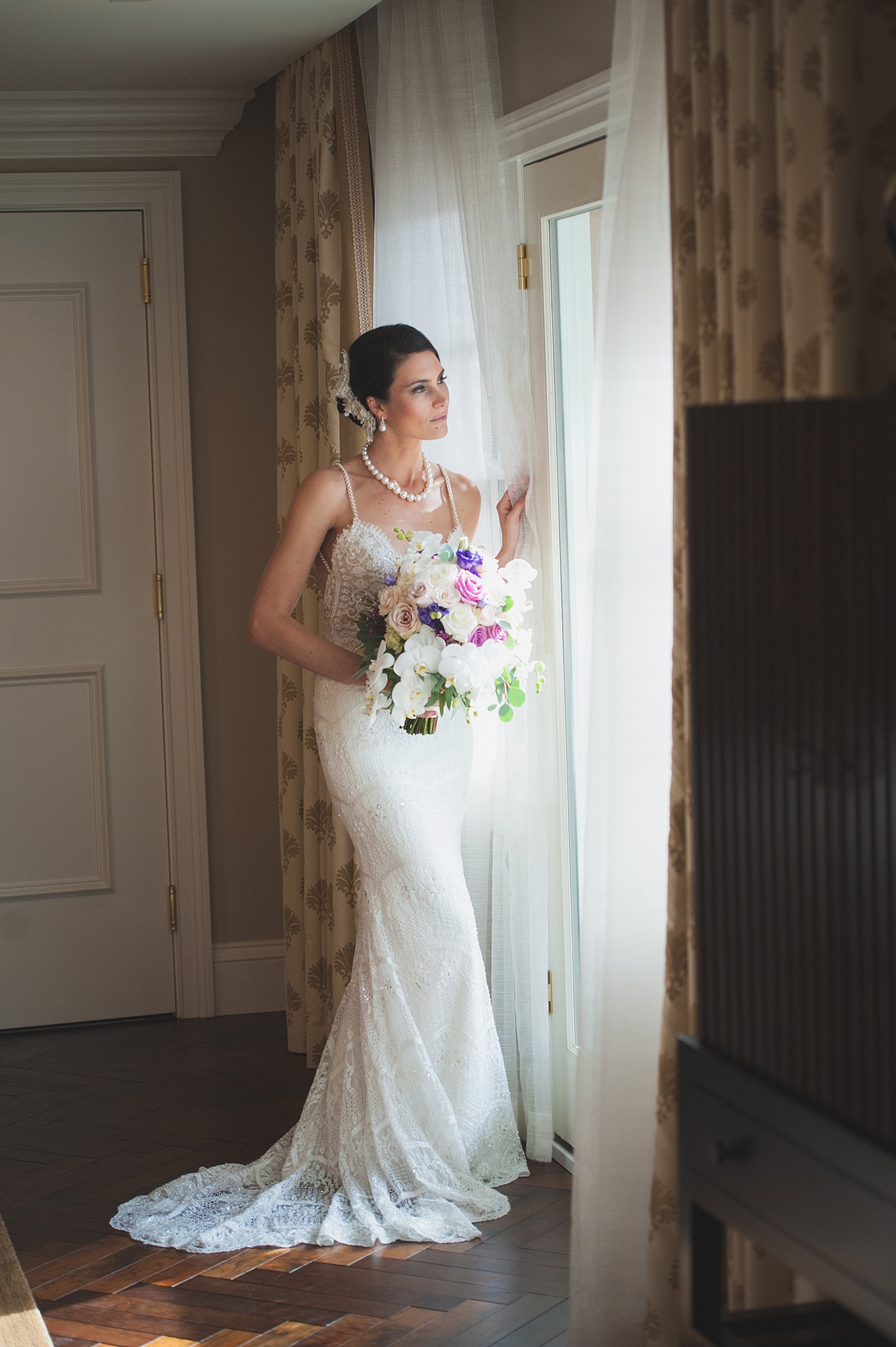 A bride stands in a door in the curtains holding her white and purple bouquet at her The Broadmoor Wedding