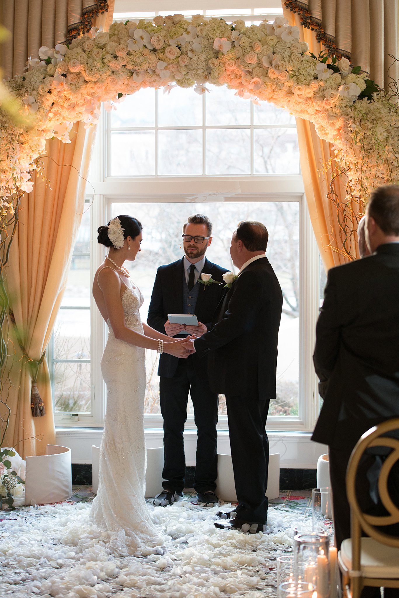 A bride and groom stand on a floor of white flowers petals under a matching arch during their ceremony at The Broadmoor Wedding venue