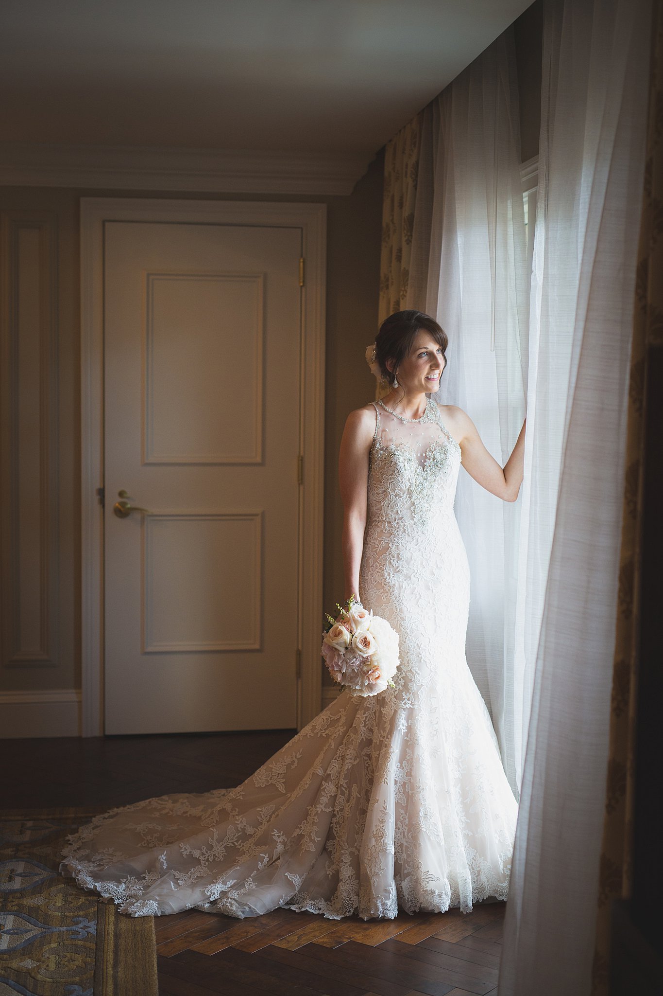 A happy bride smiles out a window while standing in the curtains holding her bouquet at her side