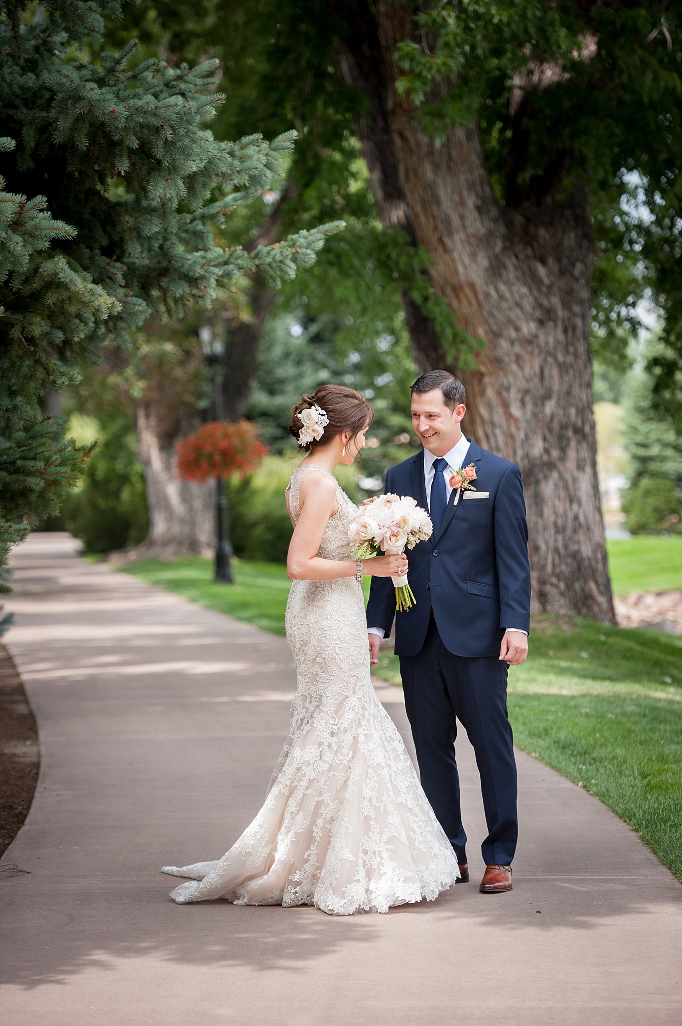 A bride and groom smile at each other under some trees on a sidewalk