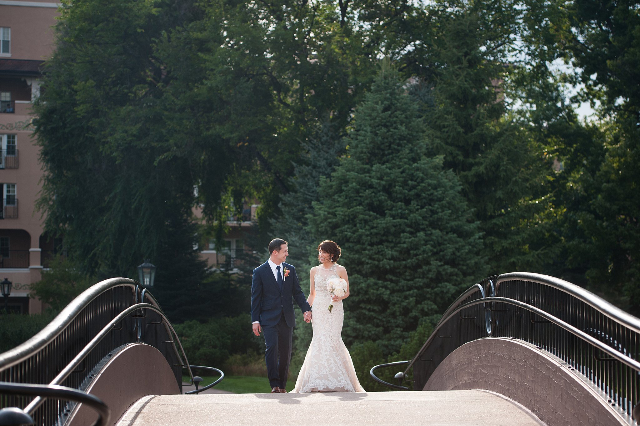 A bride and groom hold hands and smile at each other while walking over a bridge at The Broadmoor Wedding venue