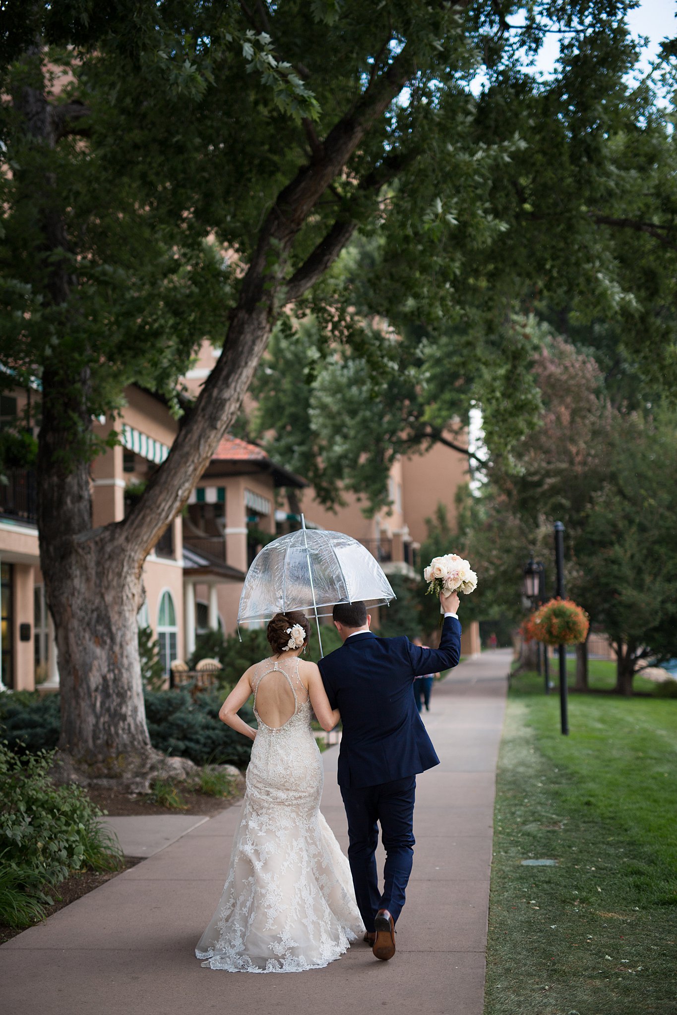 A groom lifts the bouquet up as he walks with his bride under a clear umbrella on a sidewalk