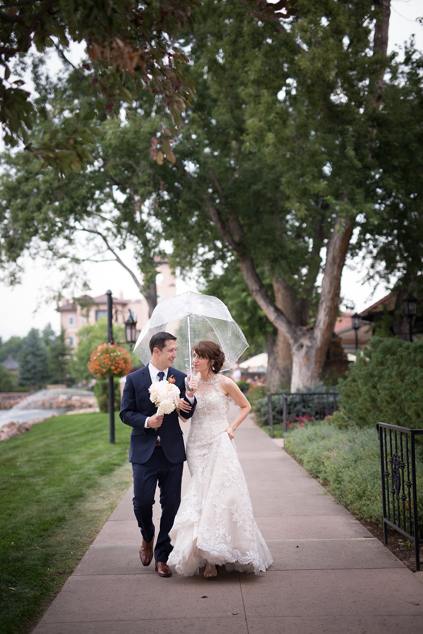 Newlyweds walk together under a clear umbrella on a sidewalk at their The Broadmoor Wedding