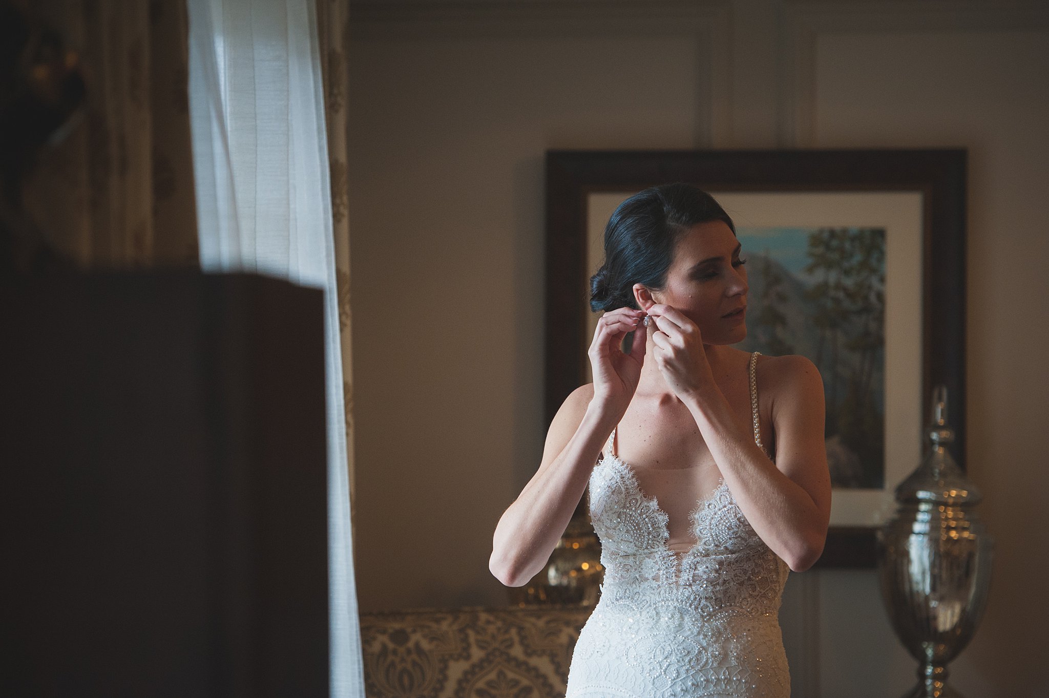 A bride in a lace embroidered dress puts on an earring while standing in the getting ready room