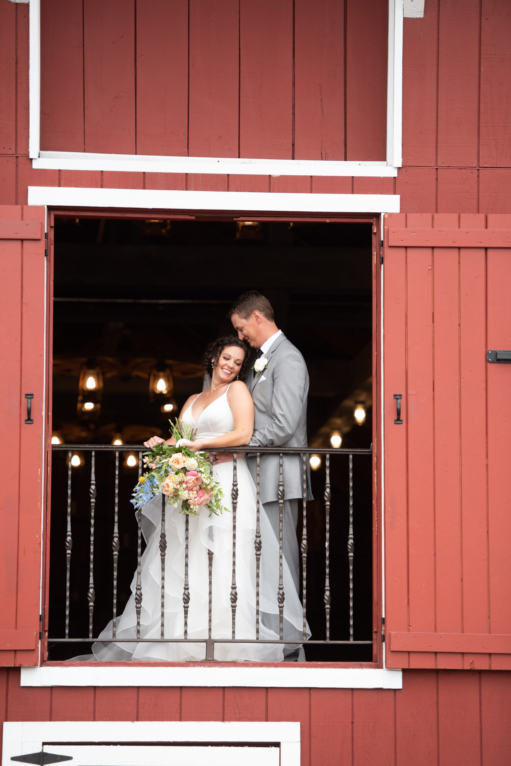 A bride and groom snuggle in a second floor window of a red barn