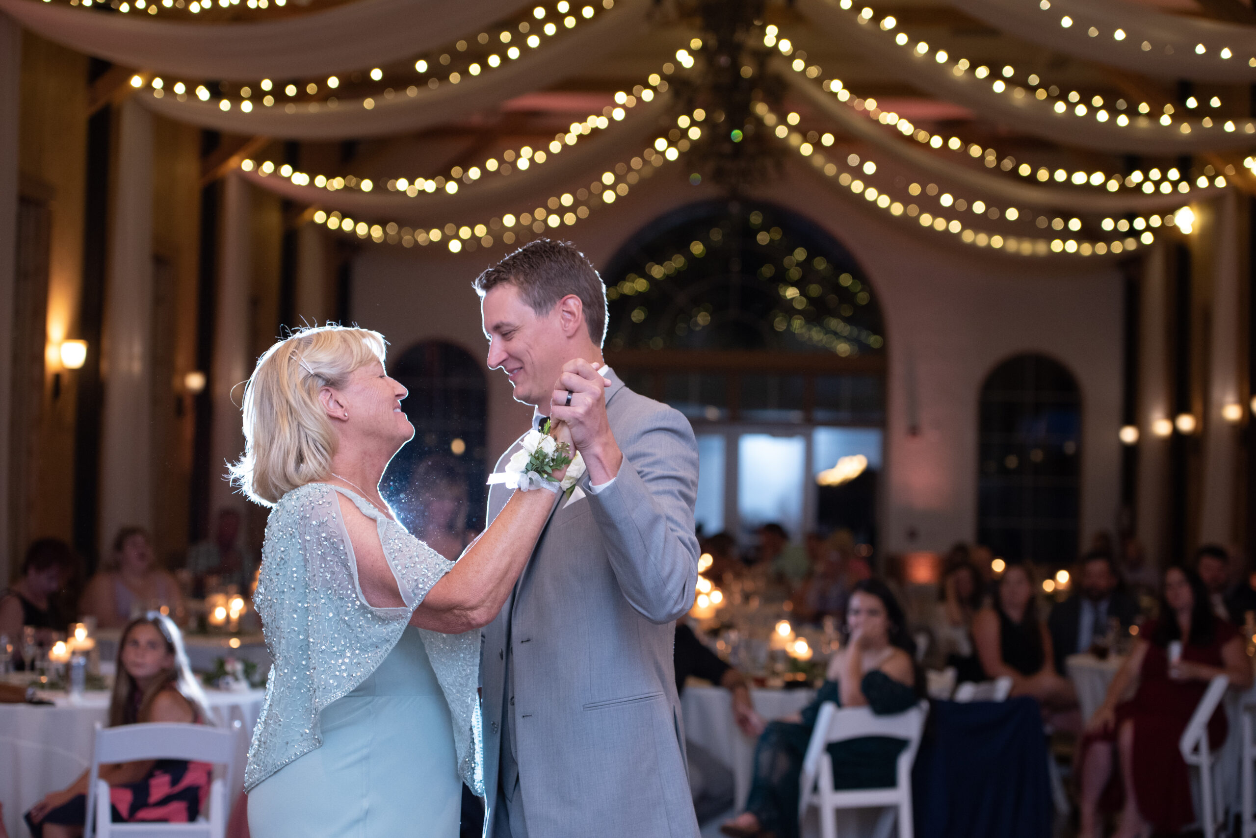 A happy mother in a blue dress sits dances with her son at his wedding under string lights