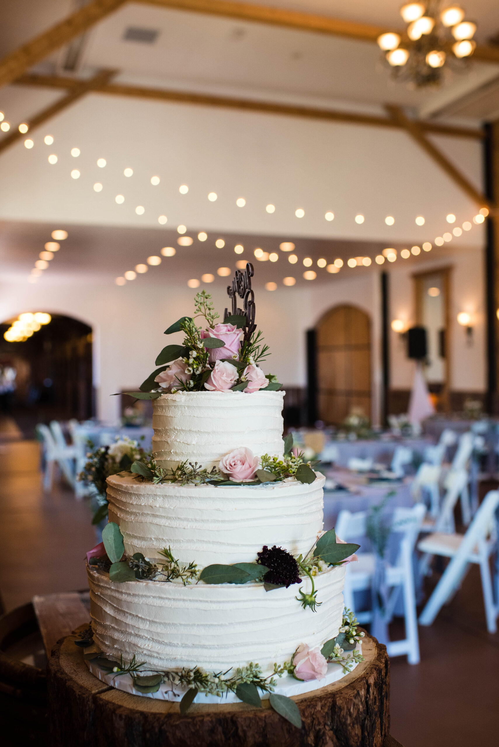 A wedding cake with pink and black flowers sits on a stump in a wedding reception