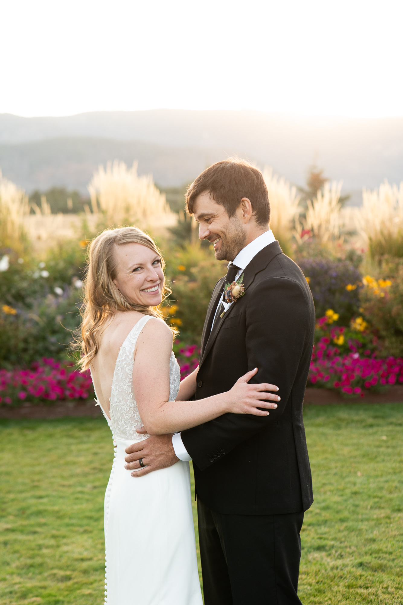 A happy bride laughs over her shoulder while hugging her groom in the colorful garden at their The Venue At Crooked Willow Farms Wedding