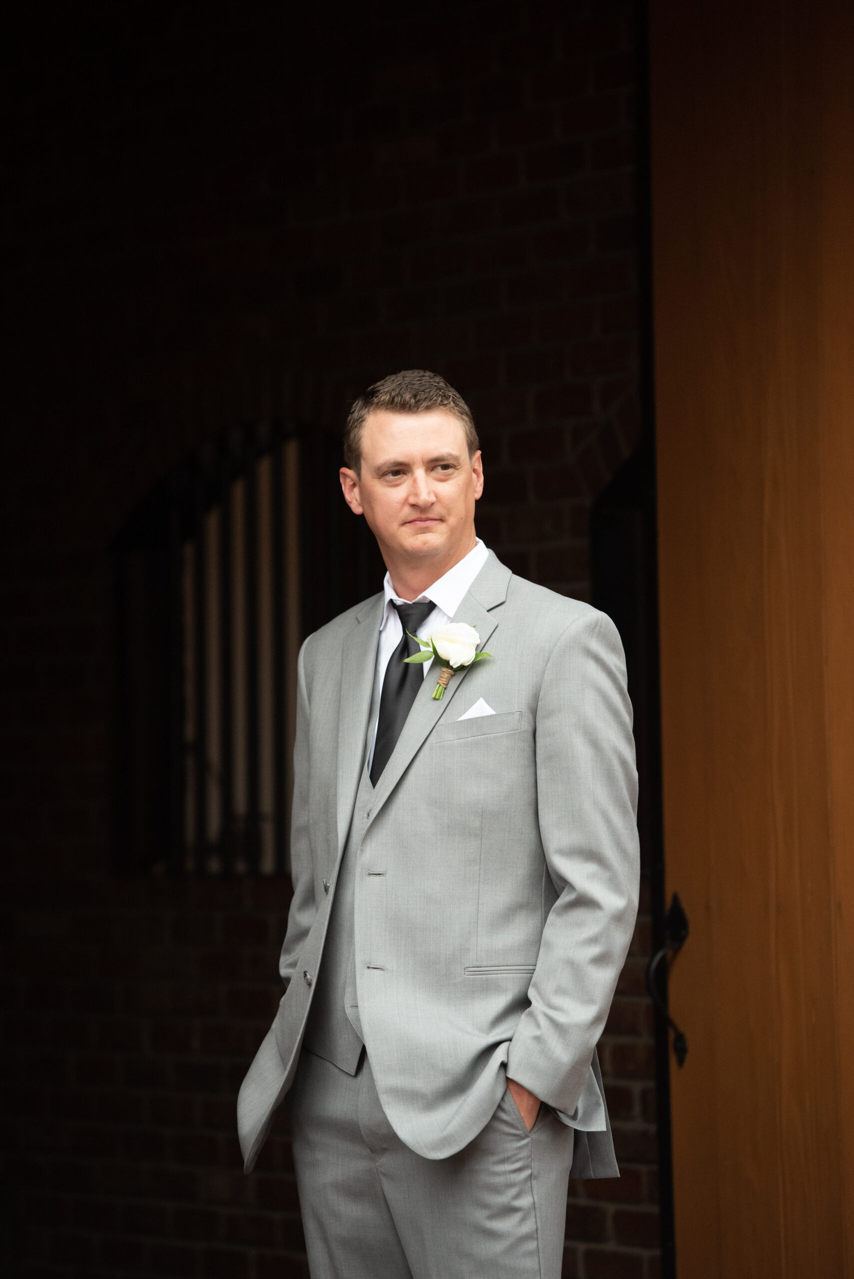 A groom stands with hands in pockets in a doorway in a grey suit