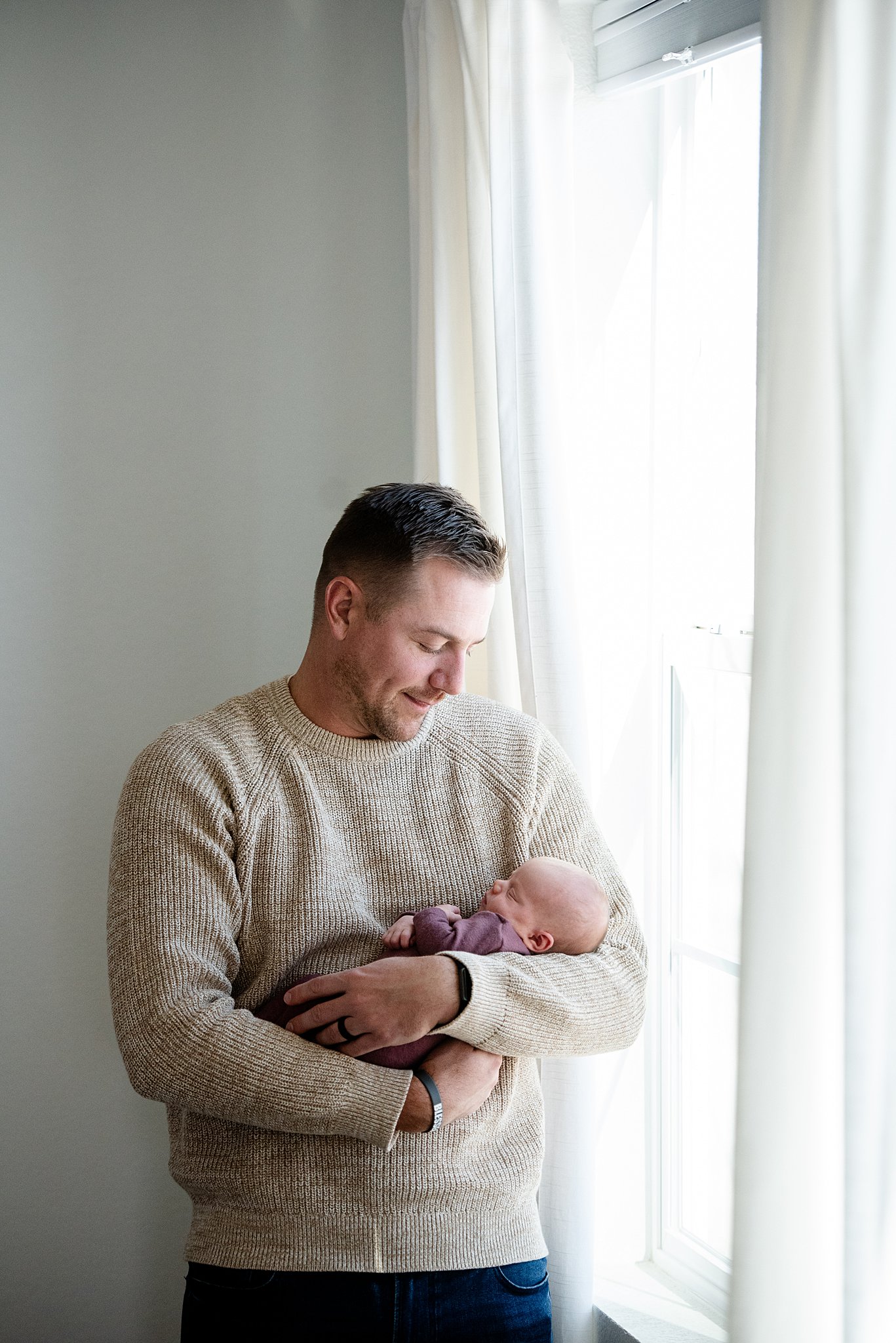 A happy new dad cradles his sleeping newborn baby in his arms while standing in a window