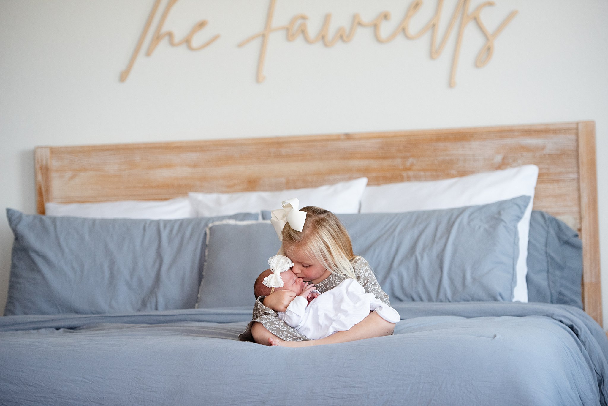 A toddler girl kisses the cheek of her newborn baby sister in her lap while sitting on a bed after visiting denver baby boutiques