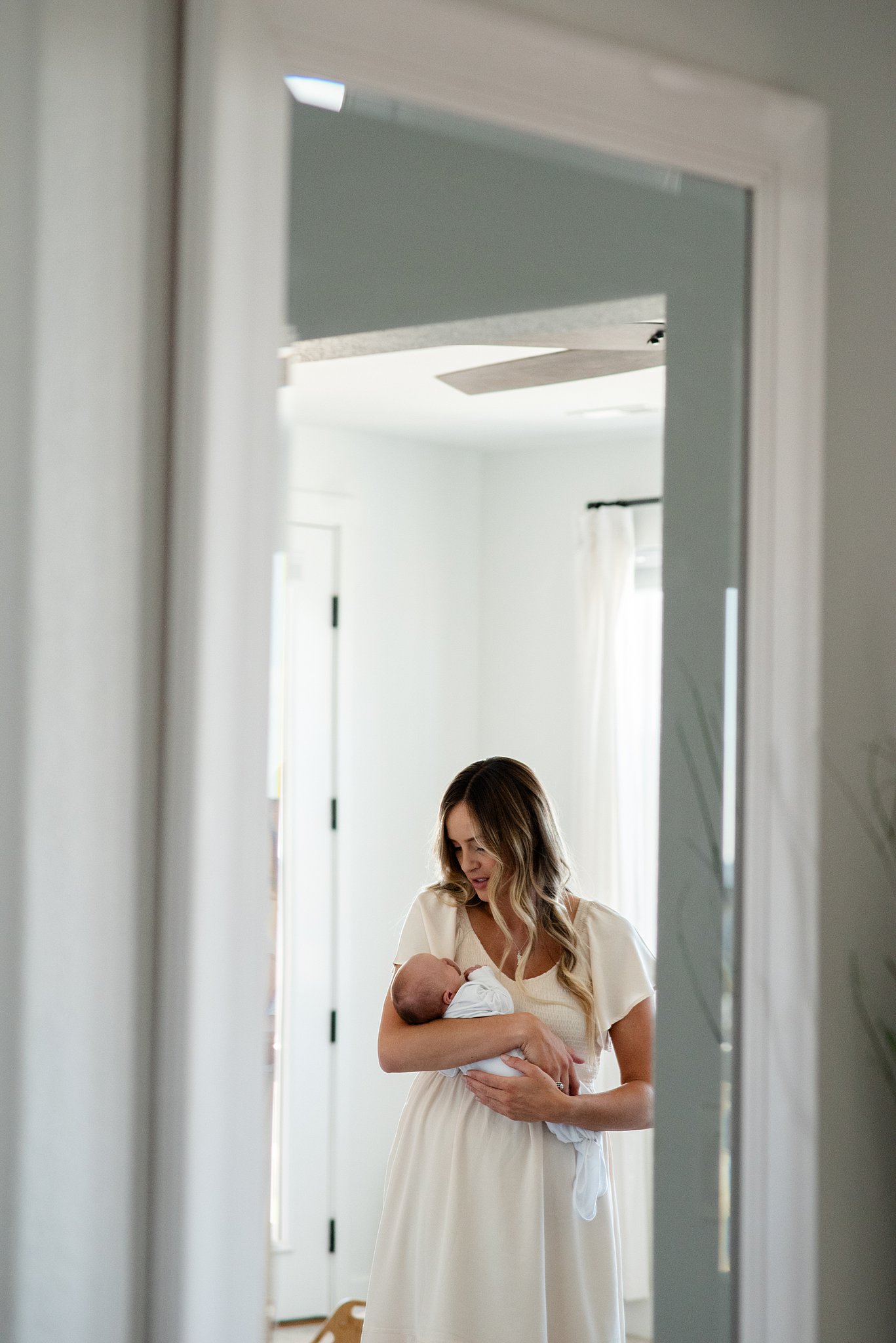 A new mom in a cream dress cradles her sleeping newborn in a mirror