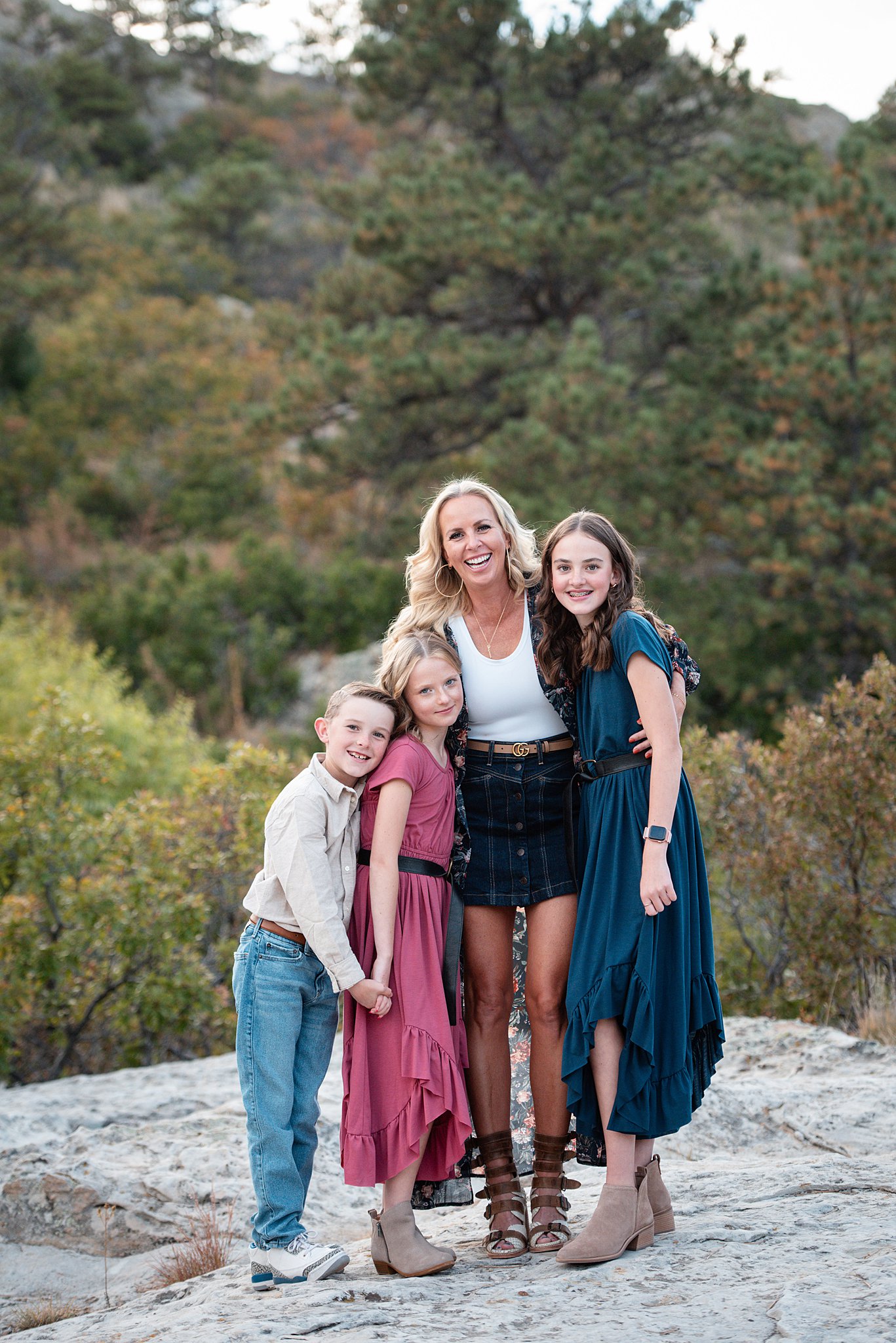 A smiling mom hugs her three young children on a boulder in dresses and jeans