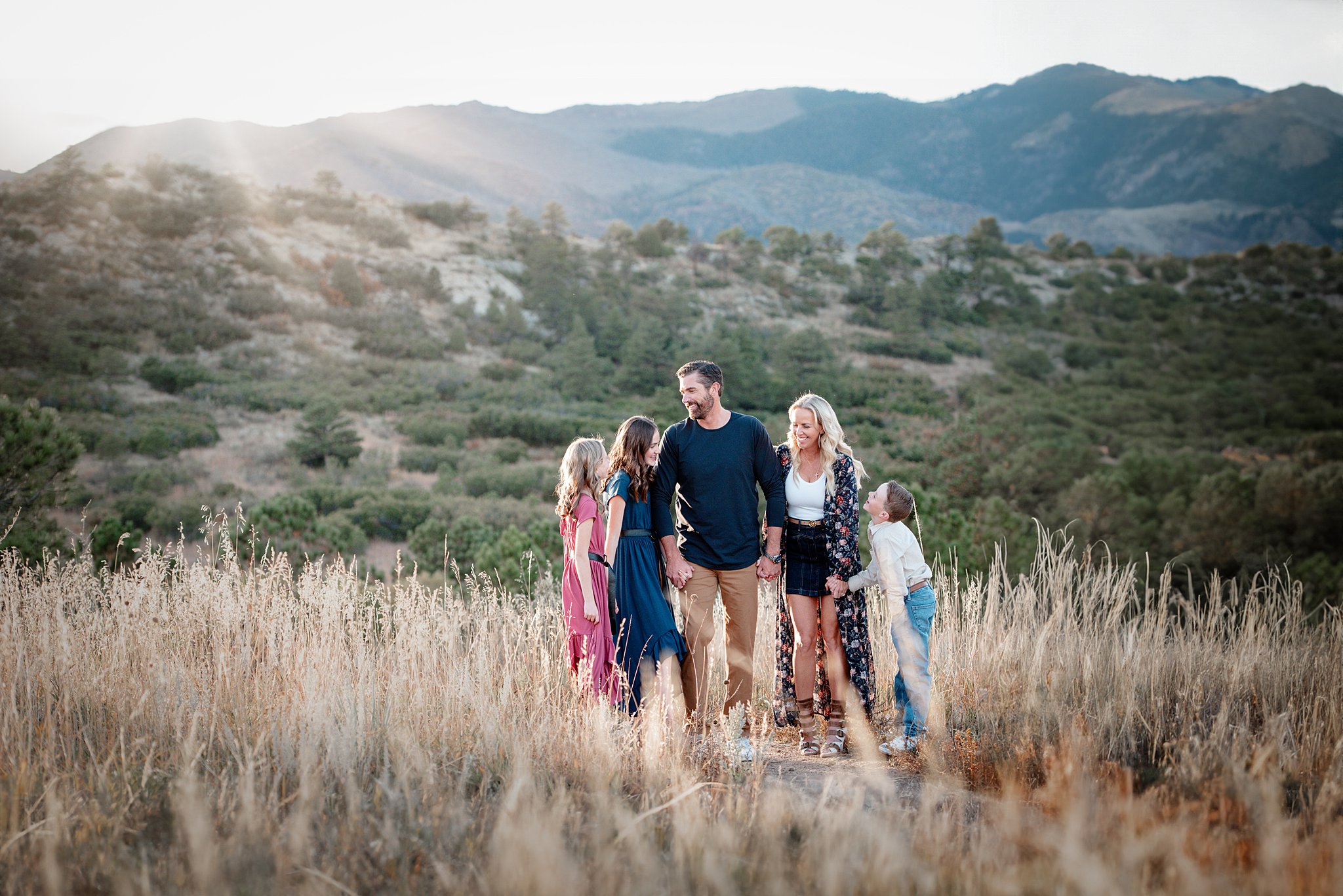 A happy family of 5 laughs while holding hands on a mountain hike at sunset after visiting Denver pediatricians