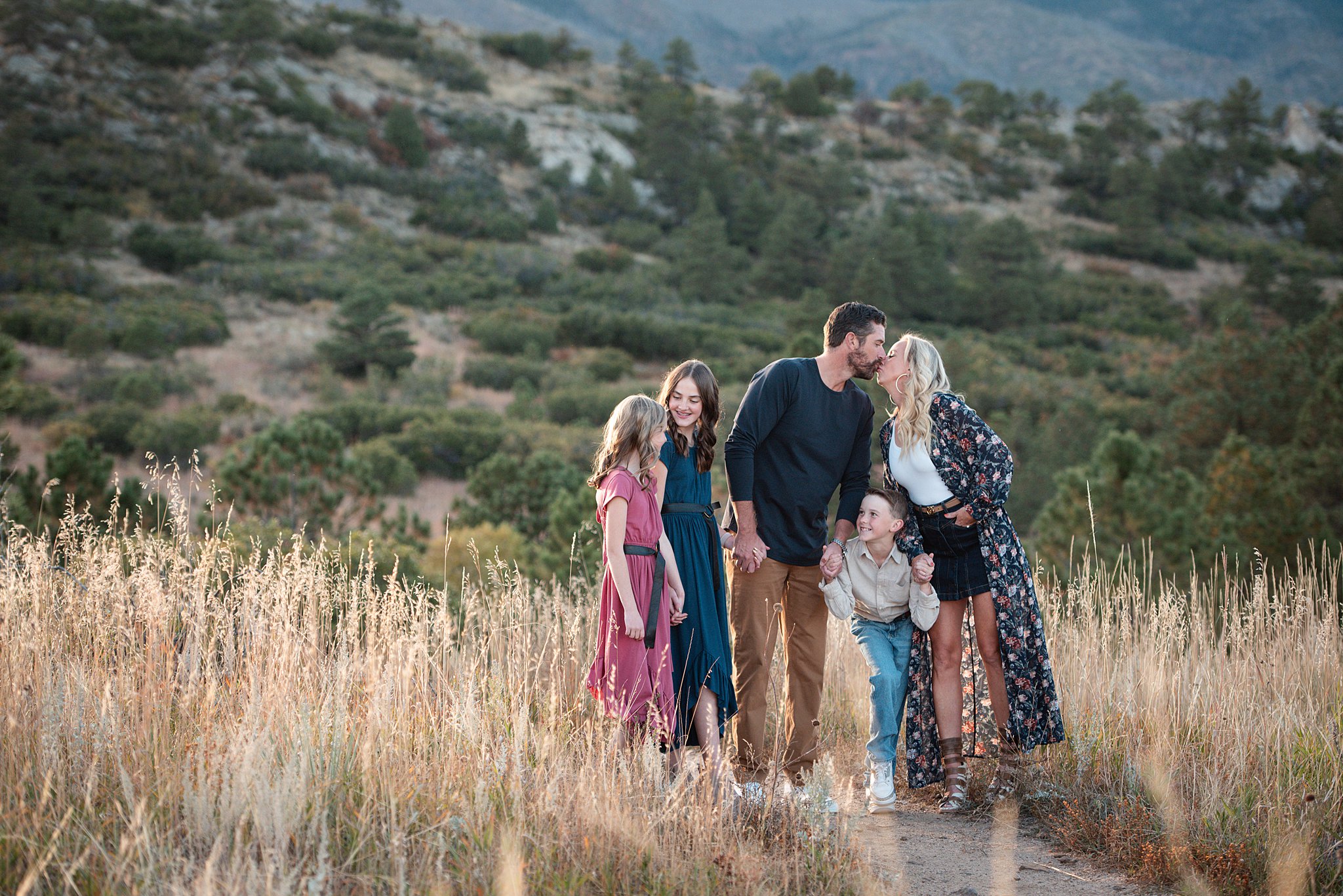 Mom and dad kiss while holding hands with their three young children on a mountain hike through tall golden grass after finding Denver pediatricians