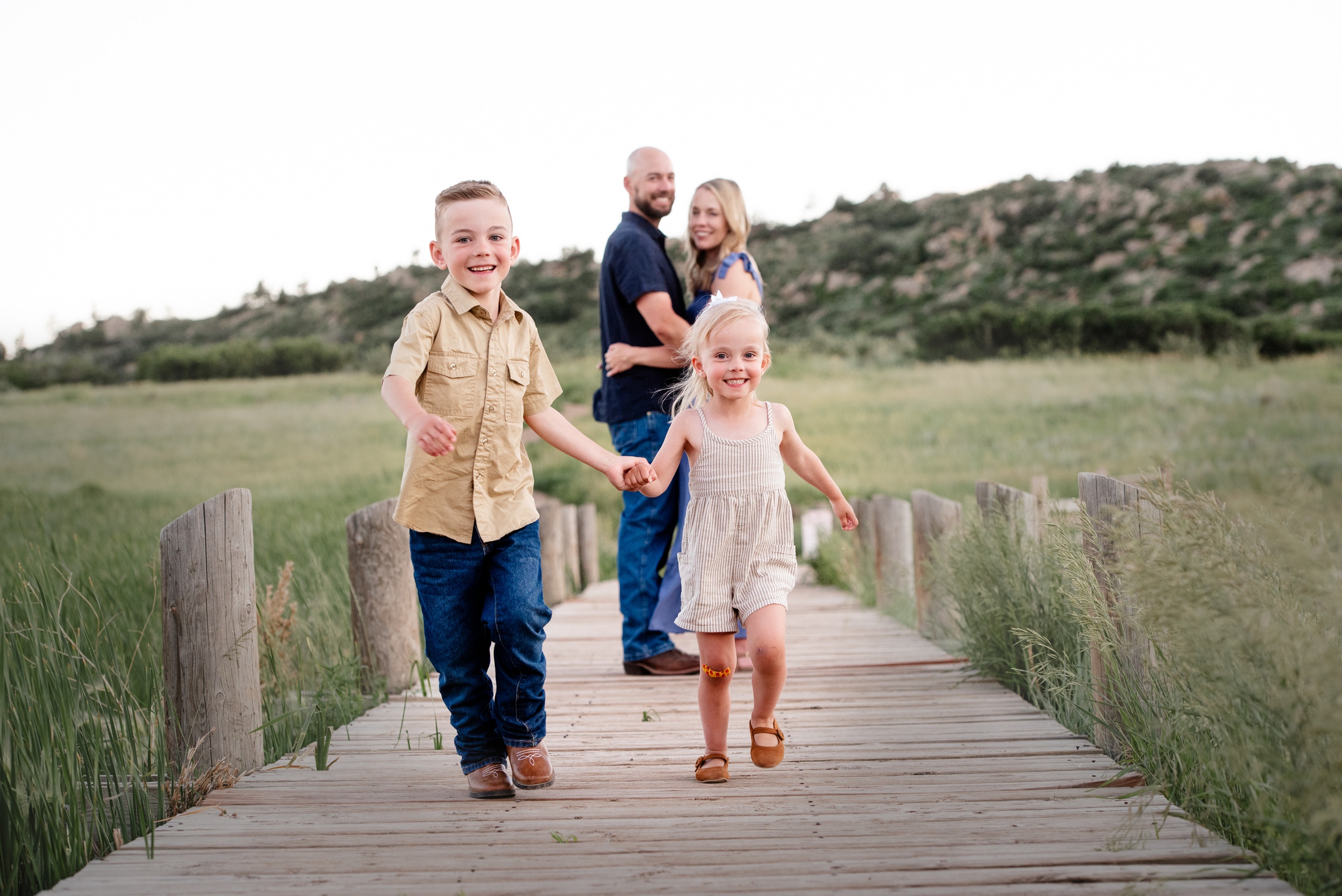 Happy toddler brother and sister run down a boardwalk as mom and dad hug behind them