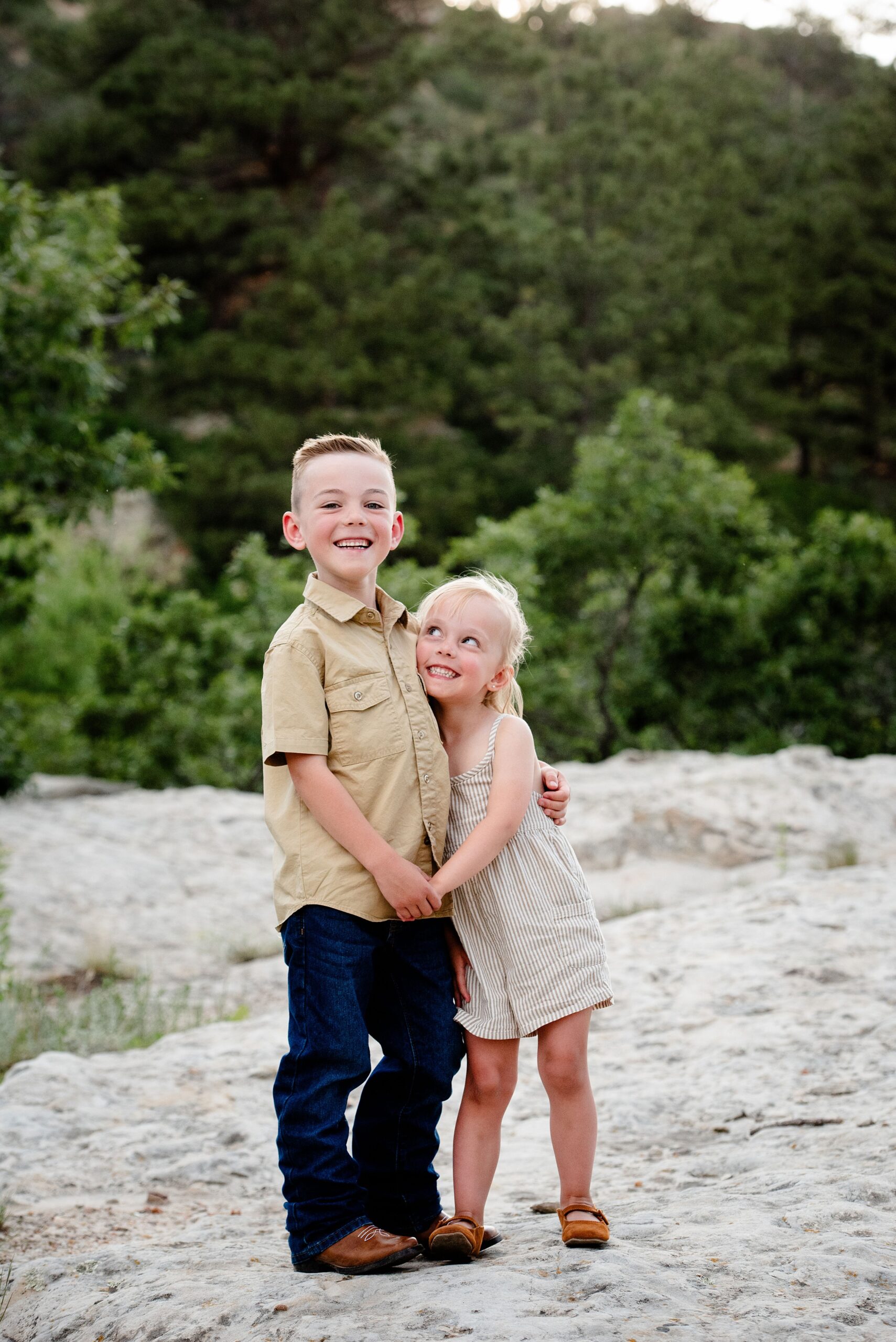 A happy toddler boy hugs his smiling baby sister while standing on a boulder at sunset after visiting denver preschools