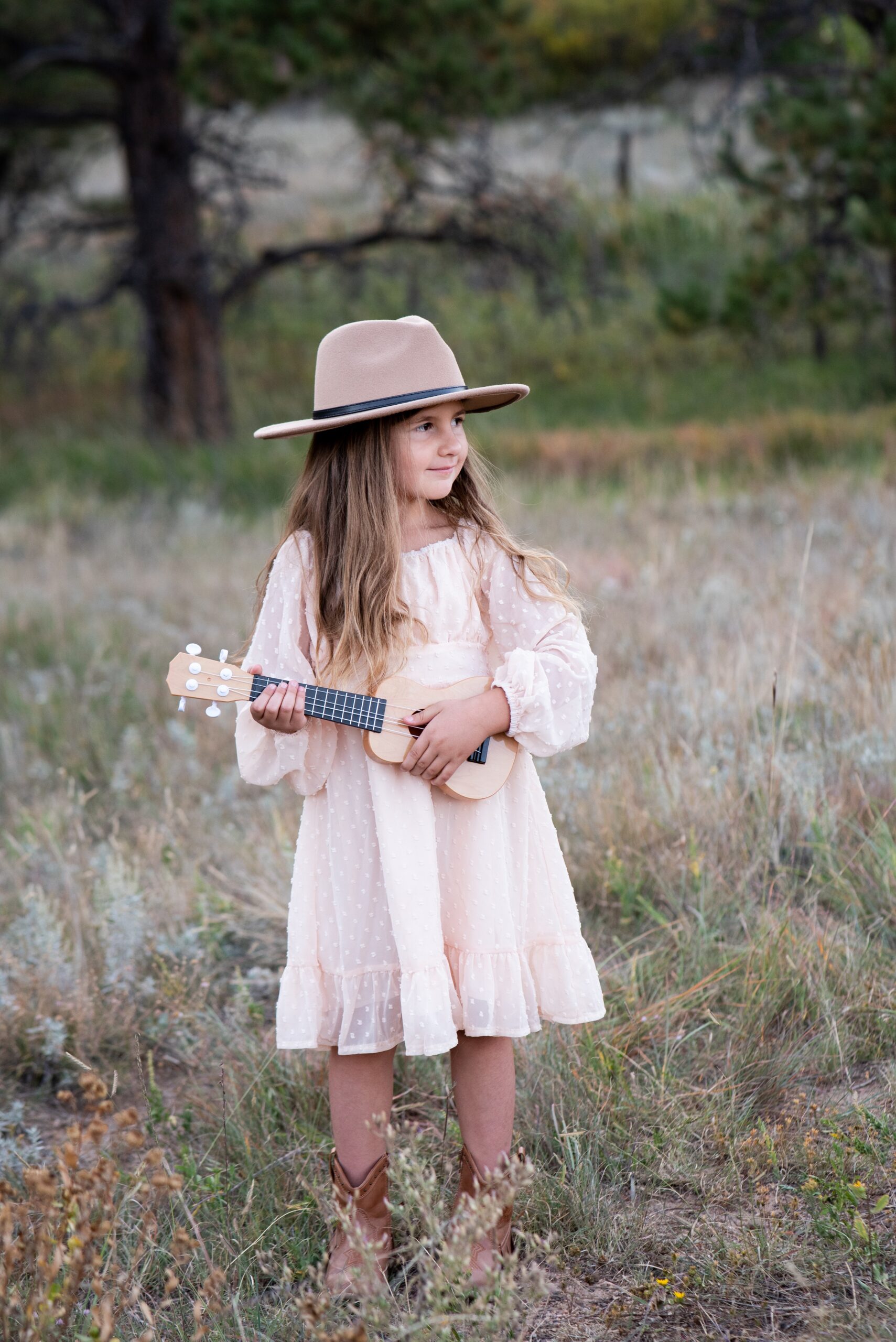 A young girl in a pink dress and brown hat plays a ukulele in a park after visiting denver toy stores