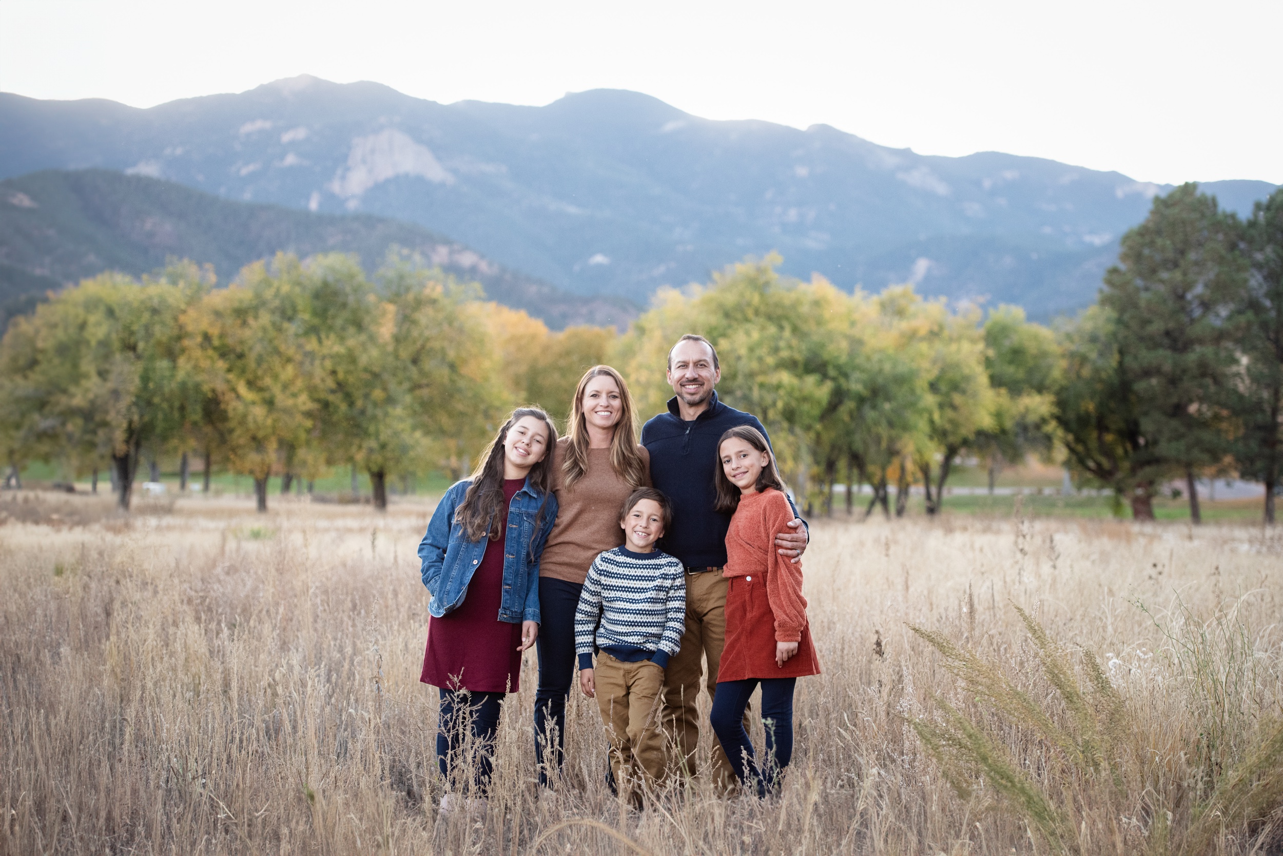 A happy family of 5 stand with arms around each other in tall golden grass after visiting indoor playgrounds in denver