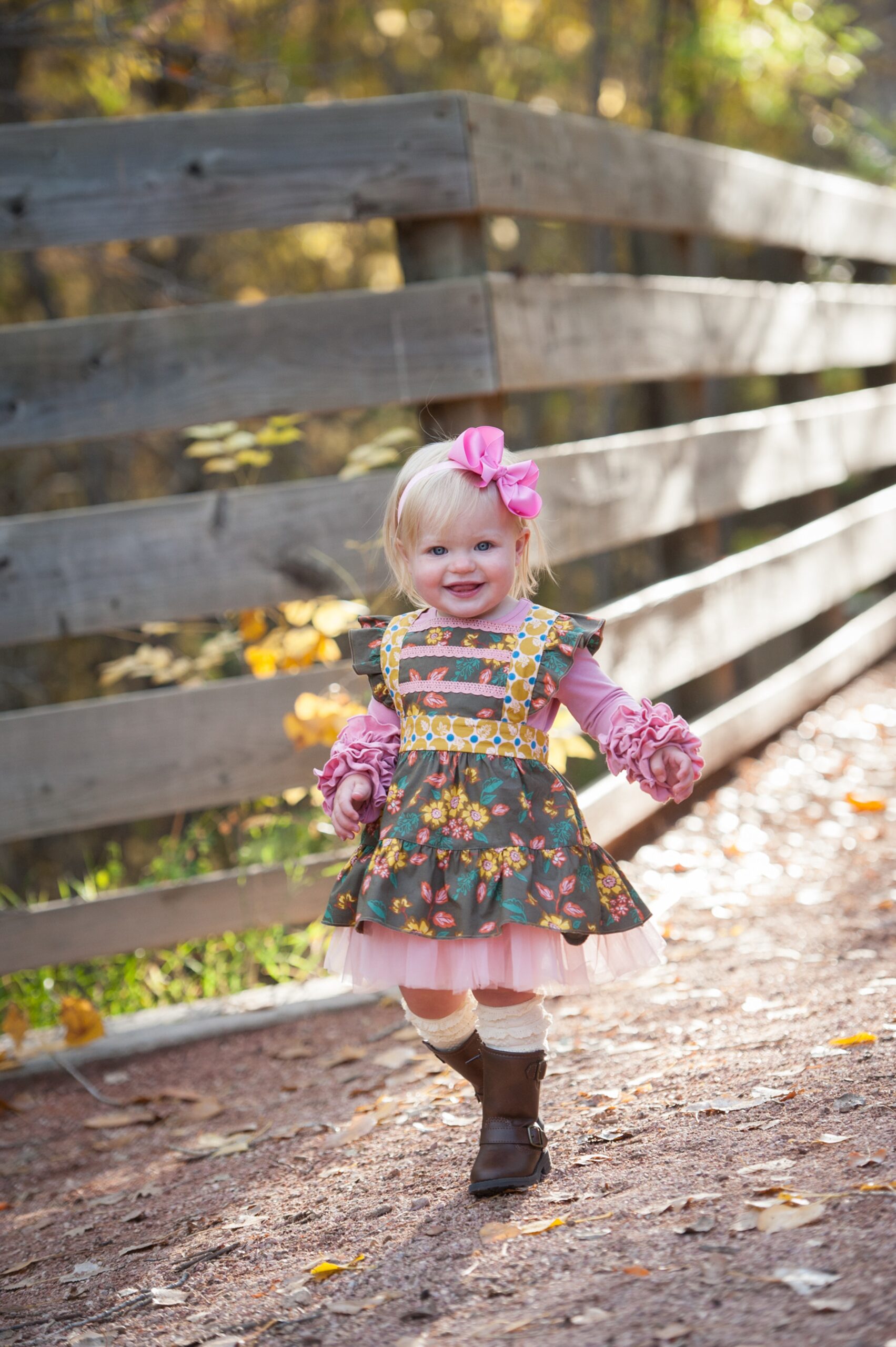 A baby girl in an elaborate pink floral dress and bow walks in a park while giggling