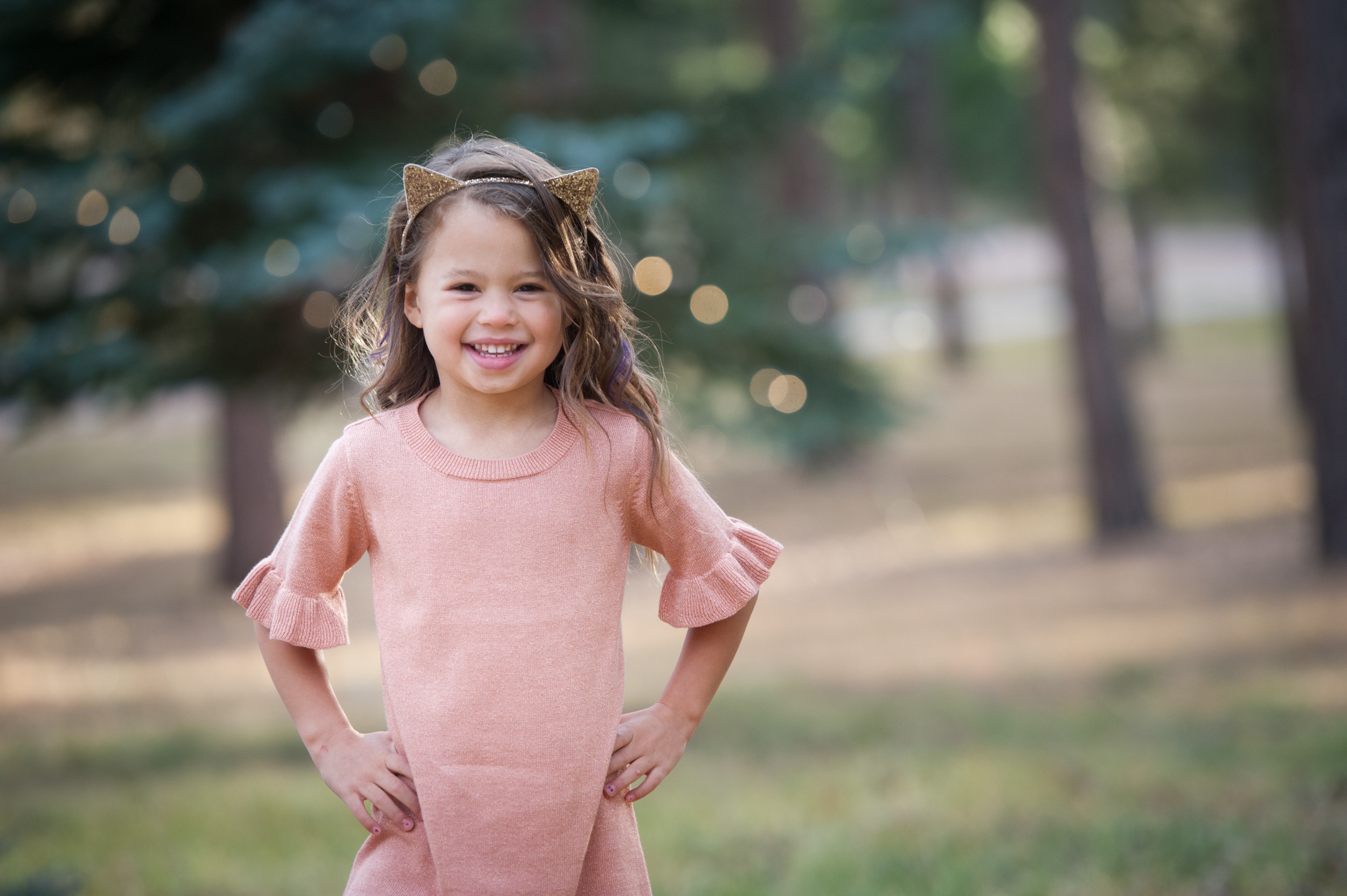 A toddler girl in a pink dress and gold cat ears stands in a park with hands on her hips smiling before visiting kid friendly restaurants in Denver