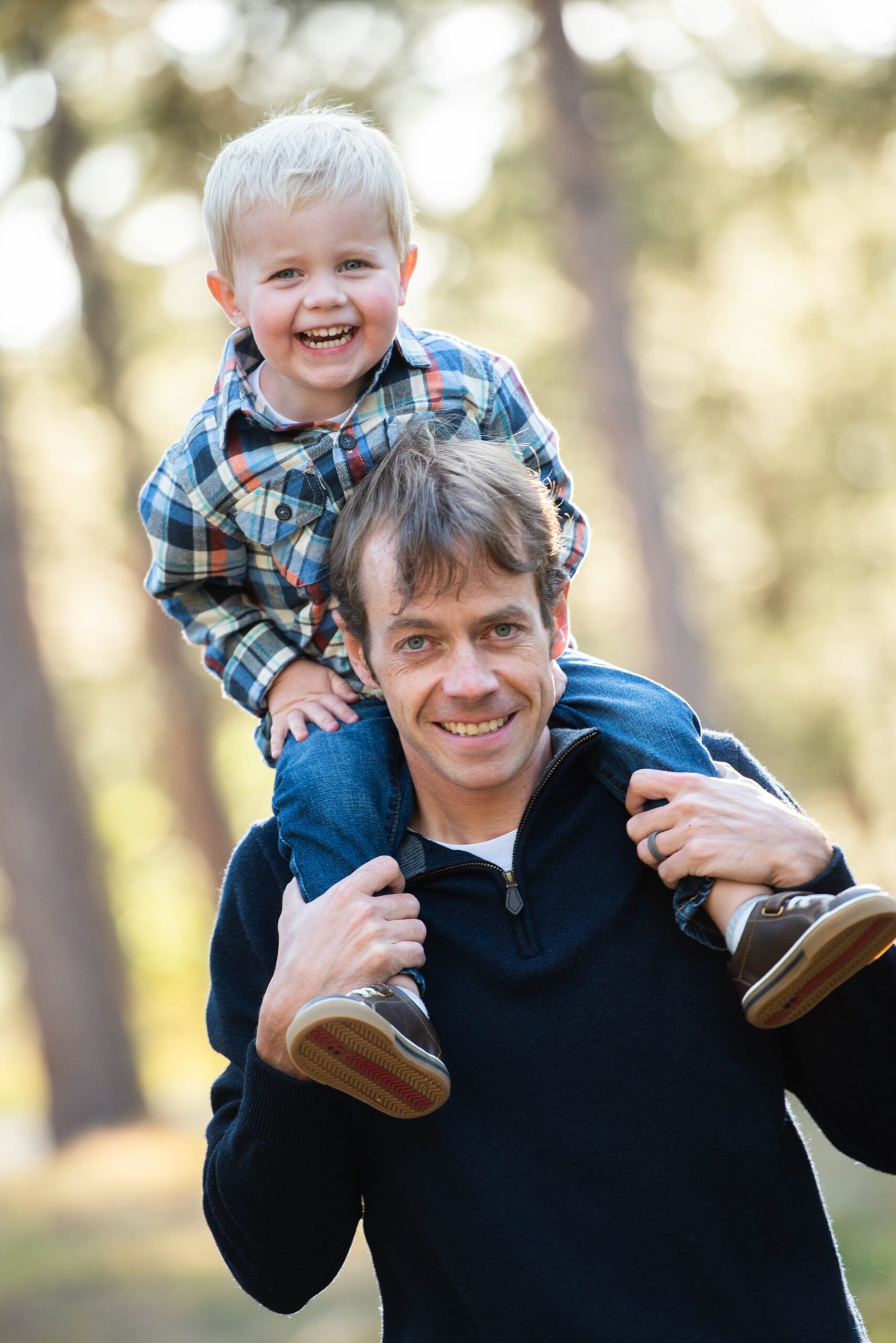 A giggling toddler boy sits on dad's shoulders in a park in a plaid shirt before visiting kid friendly restaurants in Denver