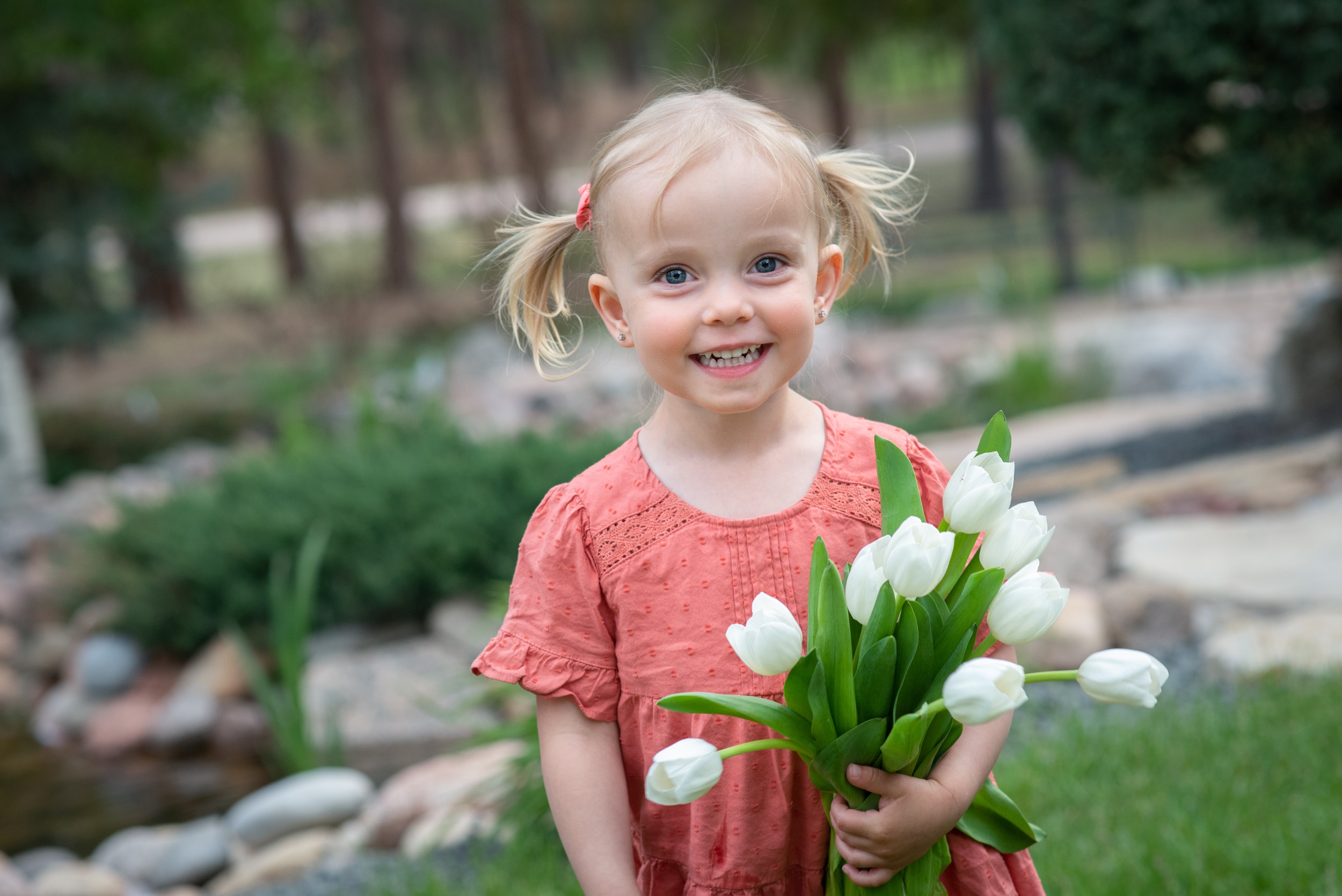 A toddler girl in a pink dress smiles big while standing in a park holding some white tulips