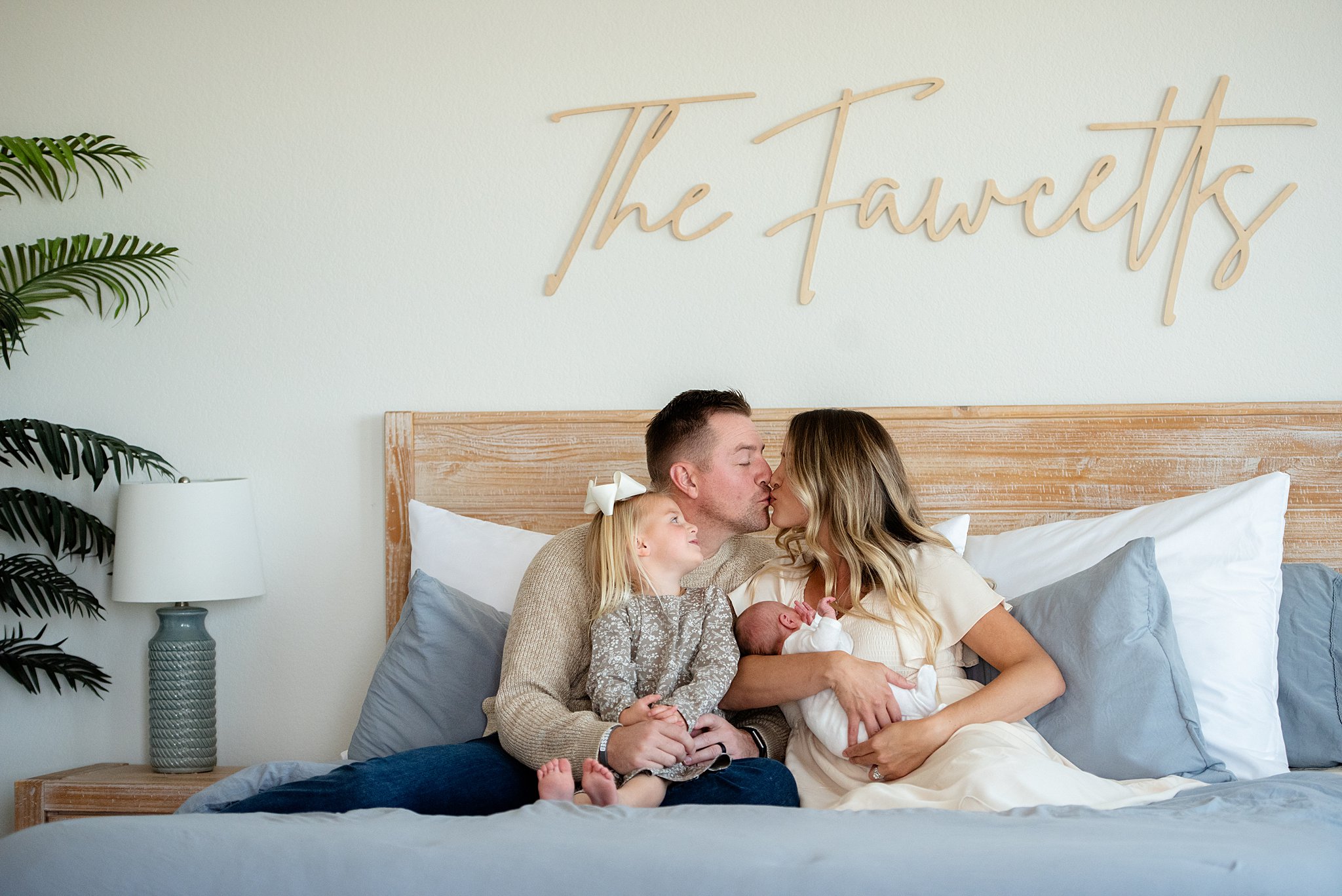 Mom and dad kiss while sitting on a bed with their newborn baby and toddler in their laps