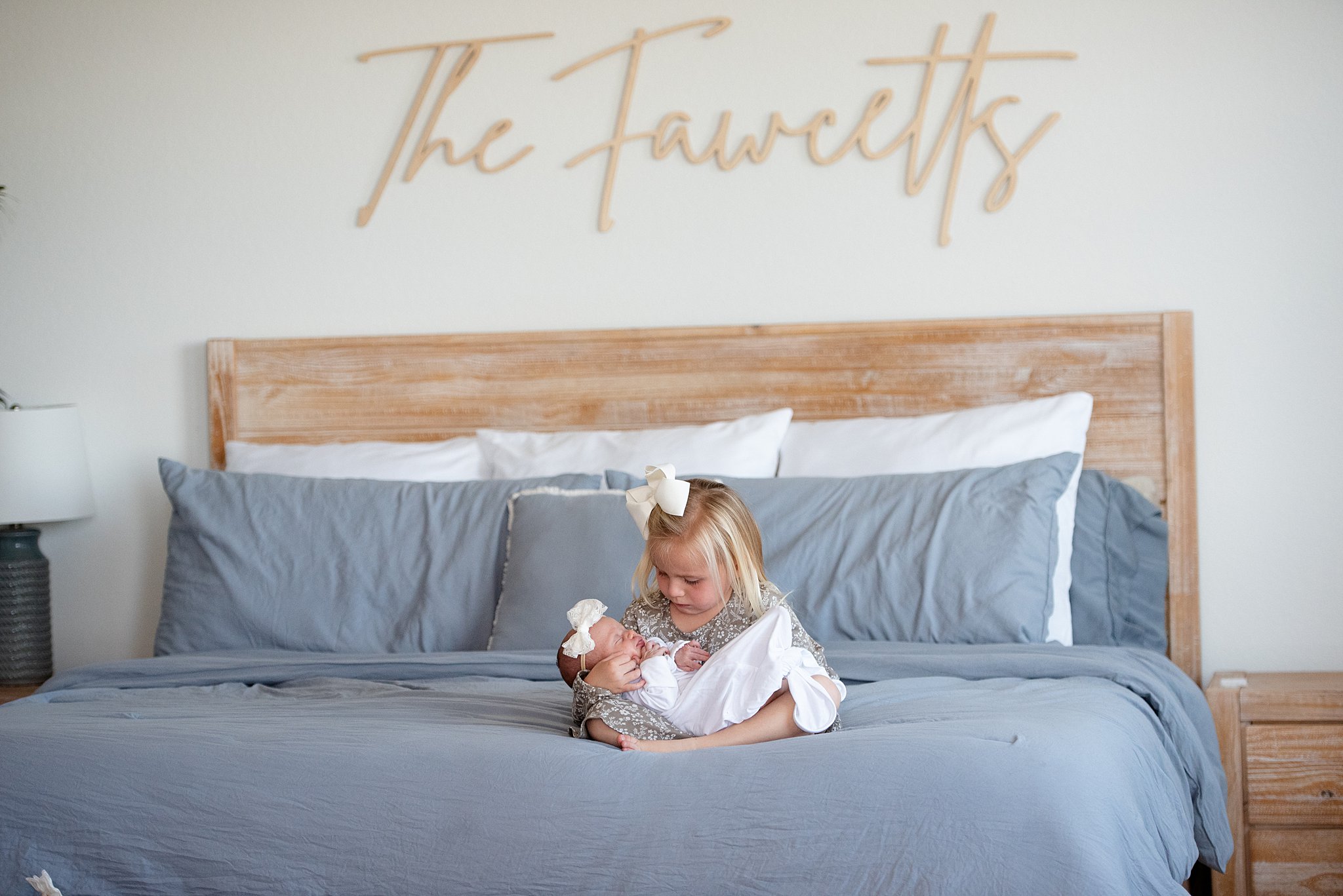 A toddler girl holds her sleeping newborn daughter in her lap on a bed in matching white bows after some mommy and me classes denver