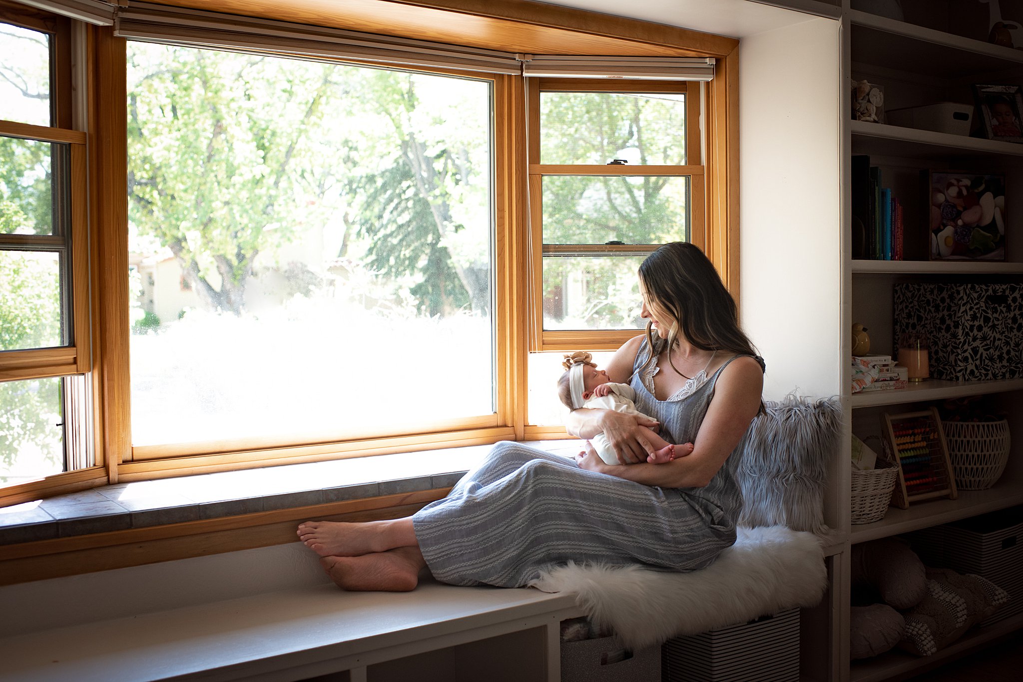 A new mom cradles her sleeping newborn daughter in a bay window shelf while in a blue dress after some mommy and me classes in denver