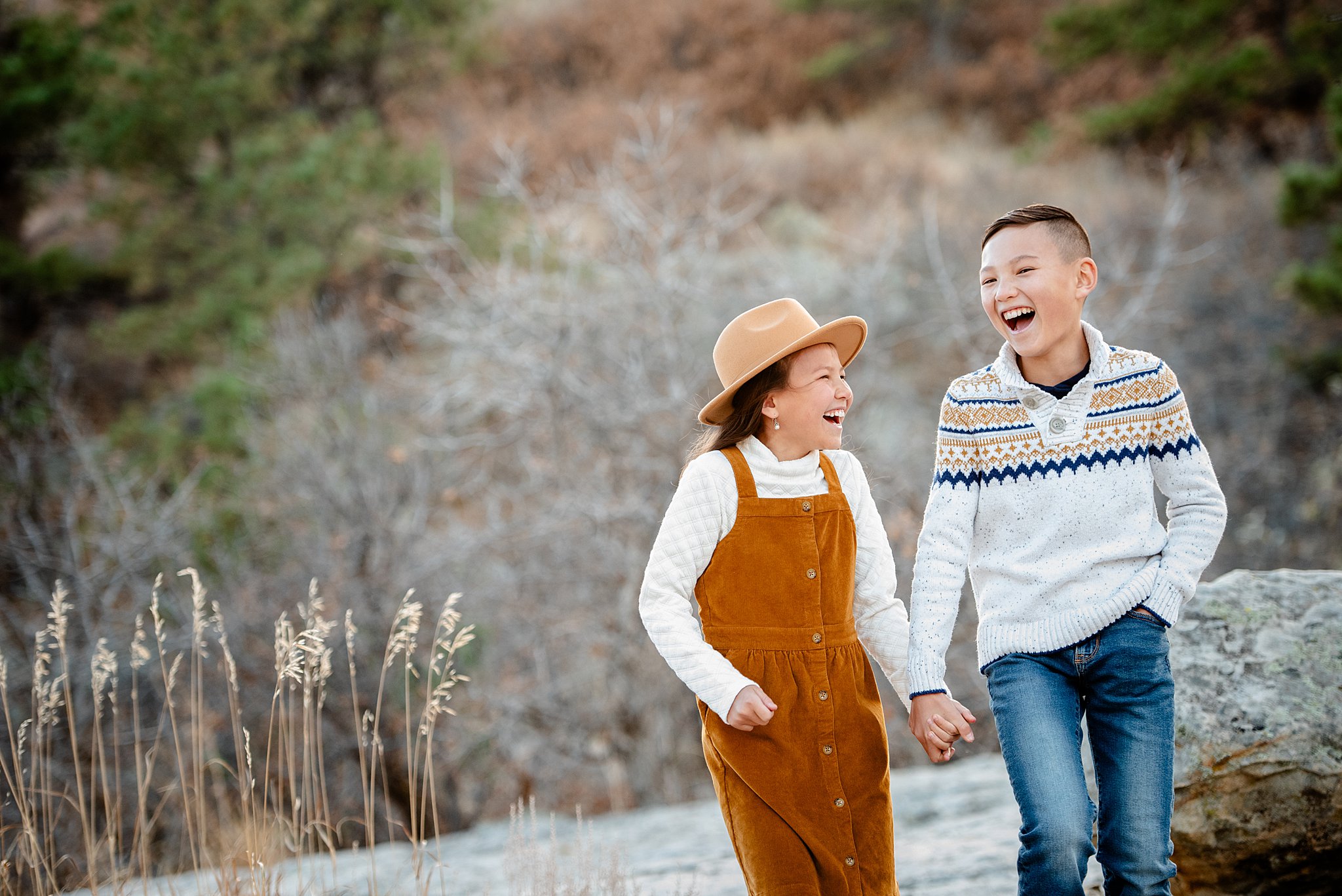 Happy brother and sister in sweaters and denim laugh while holding hands on a hike at sunset after visiting a south denver pediatric dentist