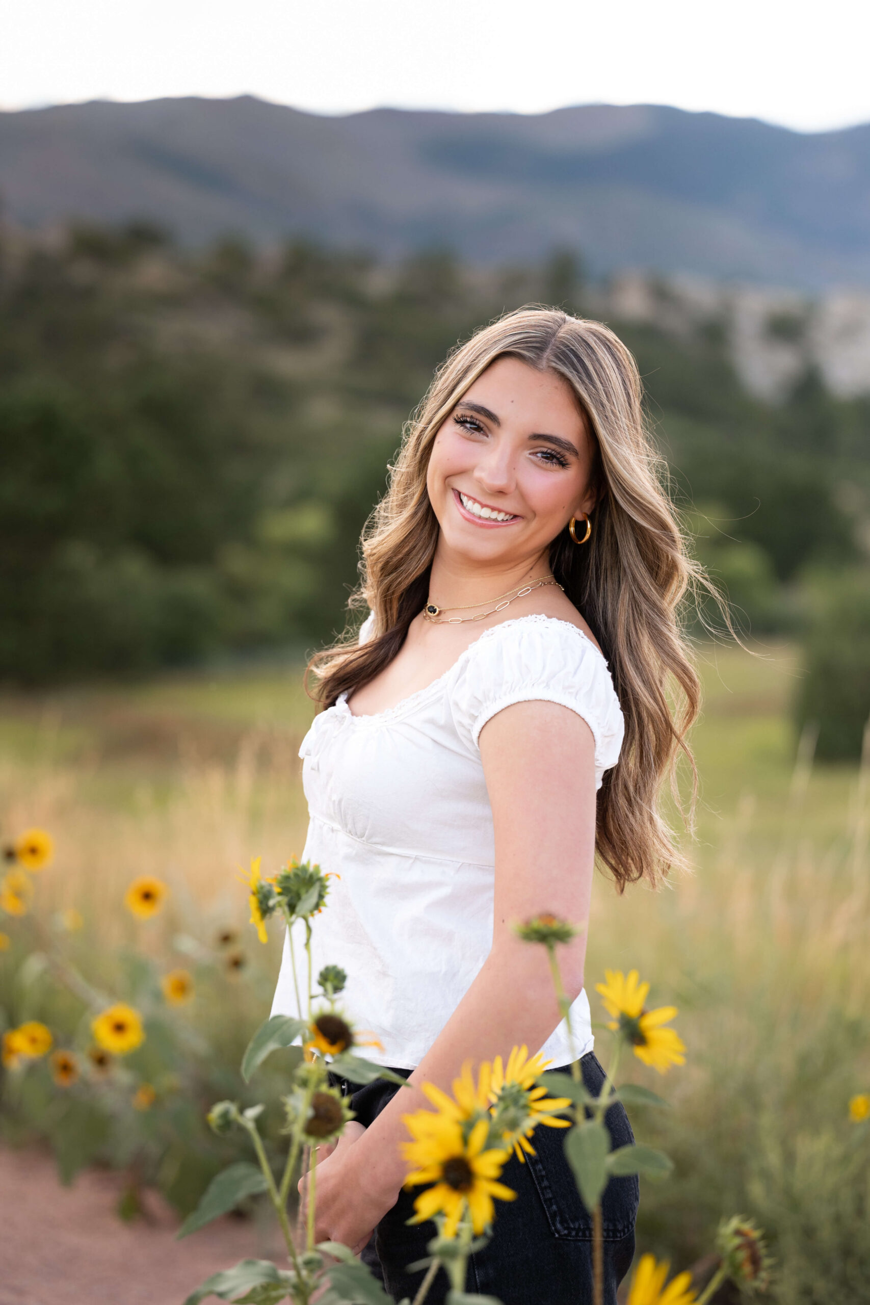 A smiling woman in a white blouse surrounded by yellow wildflowers in the mountains