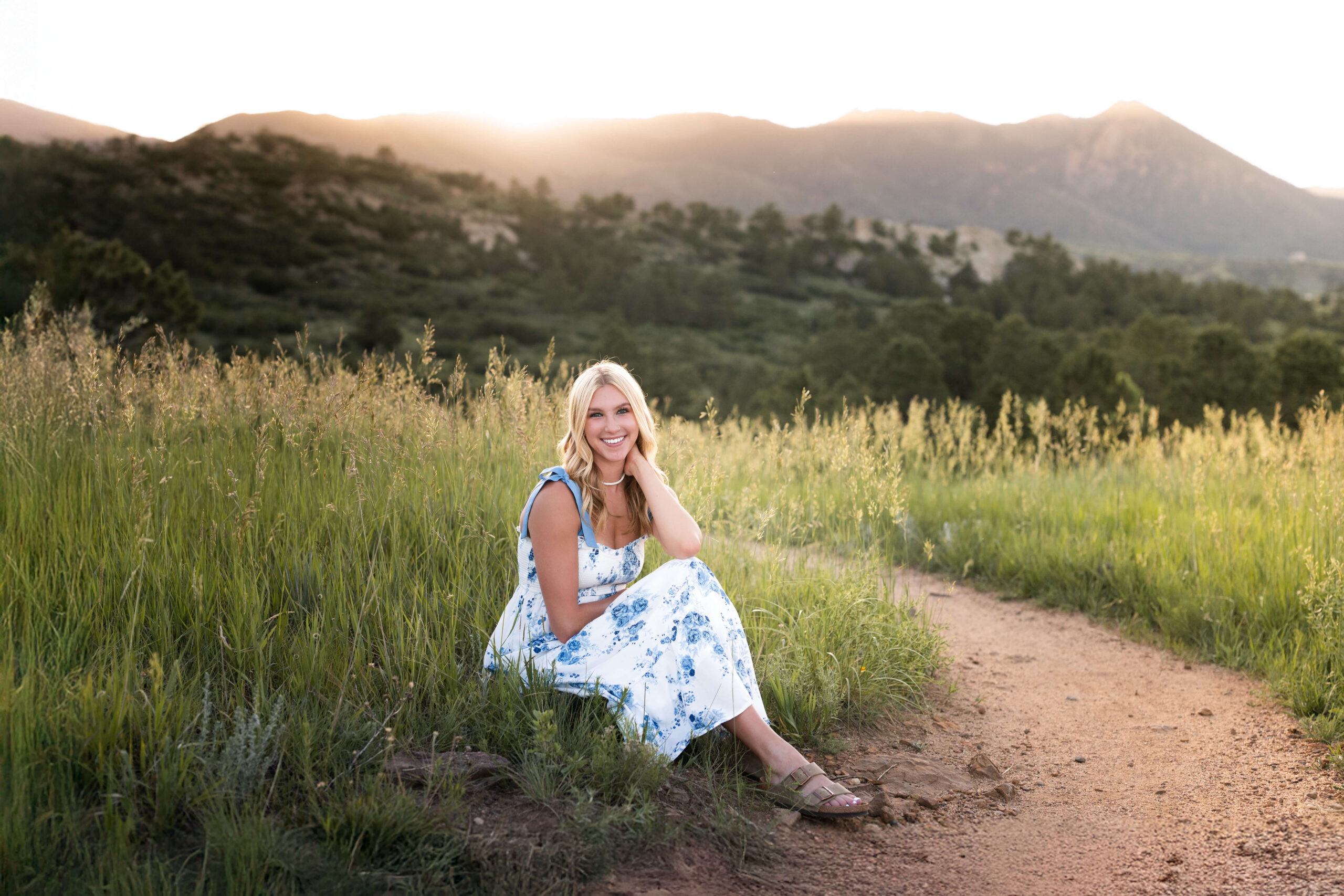 A smiling blonde graduate in a blue flower print dress sits on the edge of a mountain trail at sunset smiling after enjoying hair salons in colorado springs