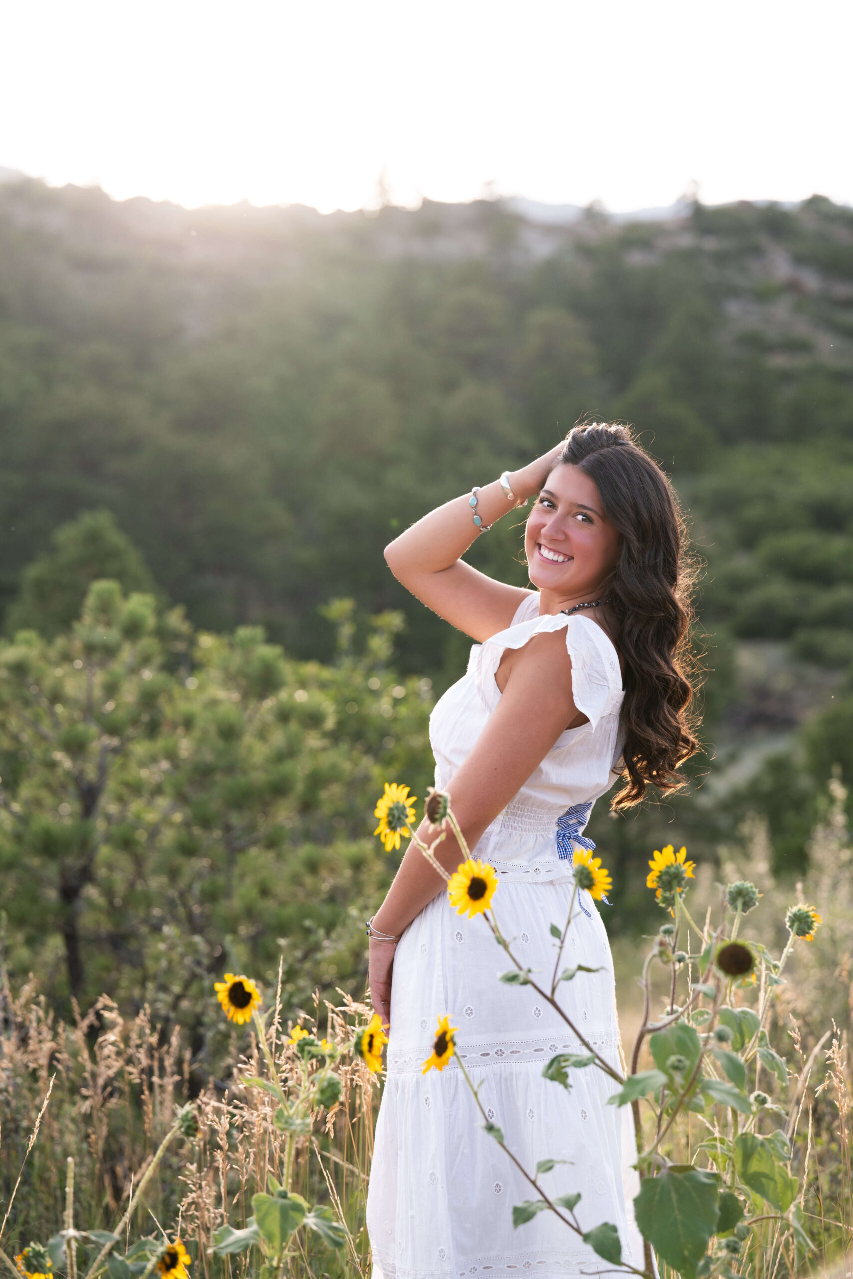 A smiling high school senior in a white dress looks over her shoulder in a mountain trail at sunset with a hand in her hair after finding hair salons in colorado springs