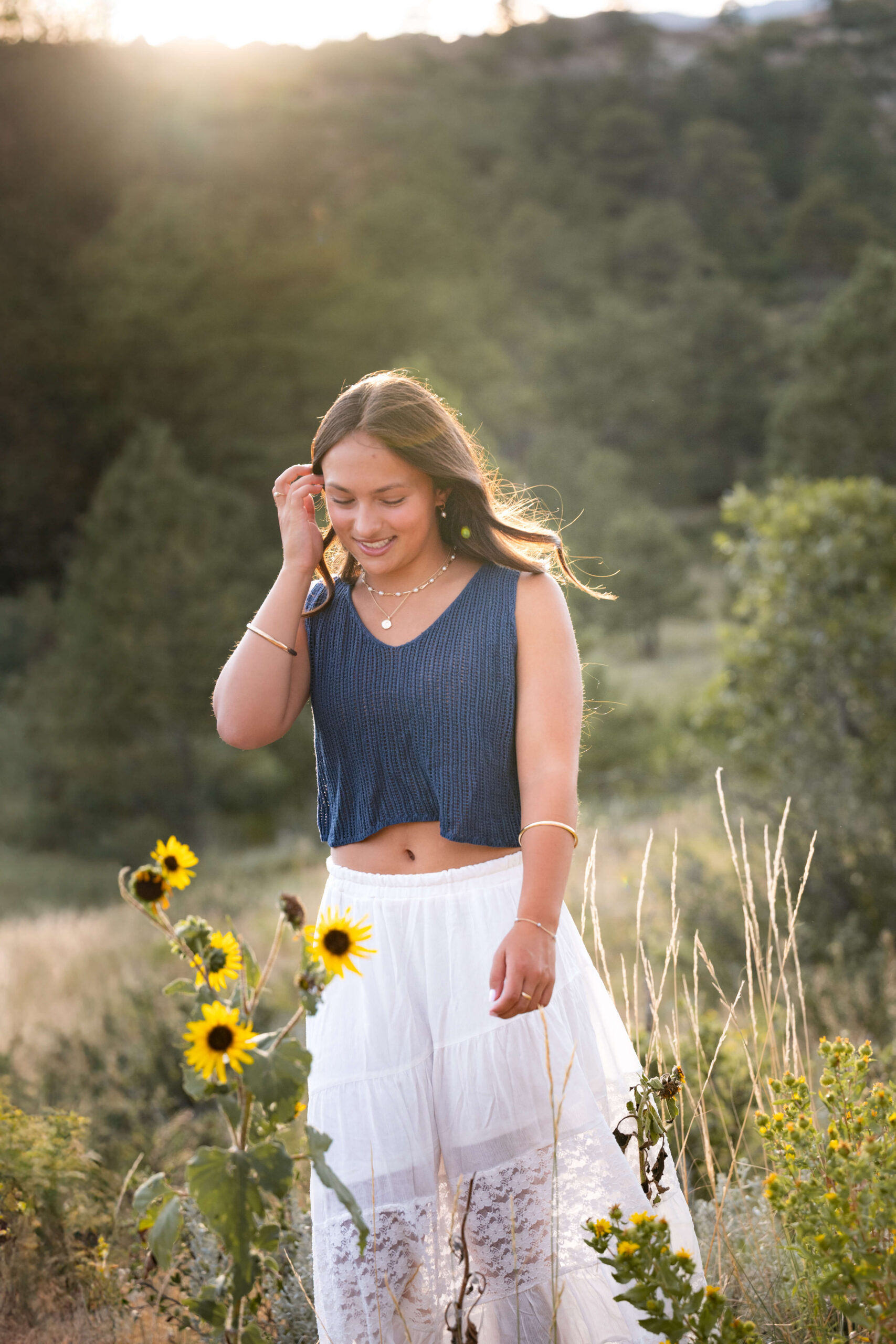 A woman pulls her hair back in the wind while walking a mountain trail with wildflowers at sunset in a blue blouse and white skirt