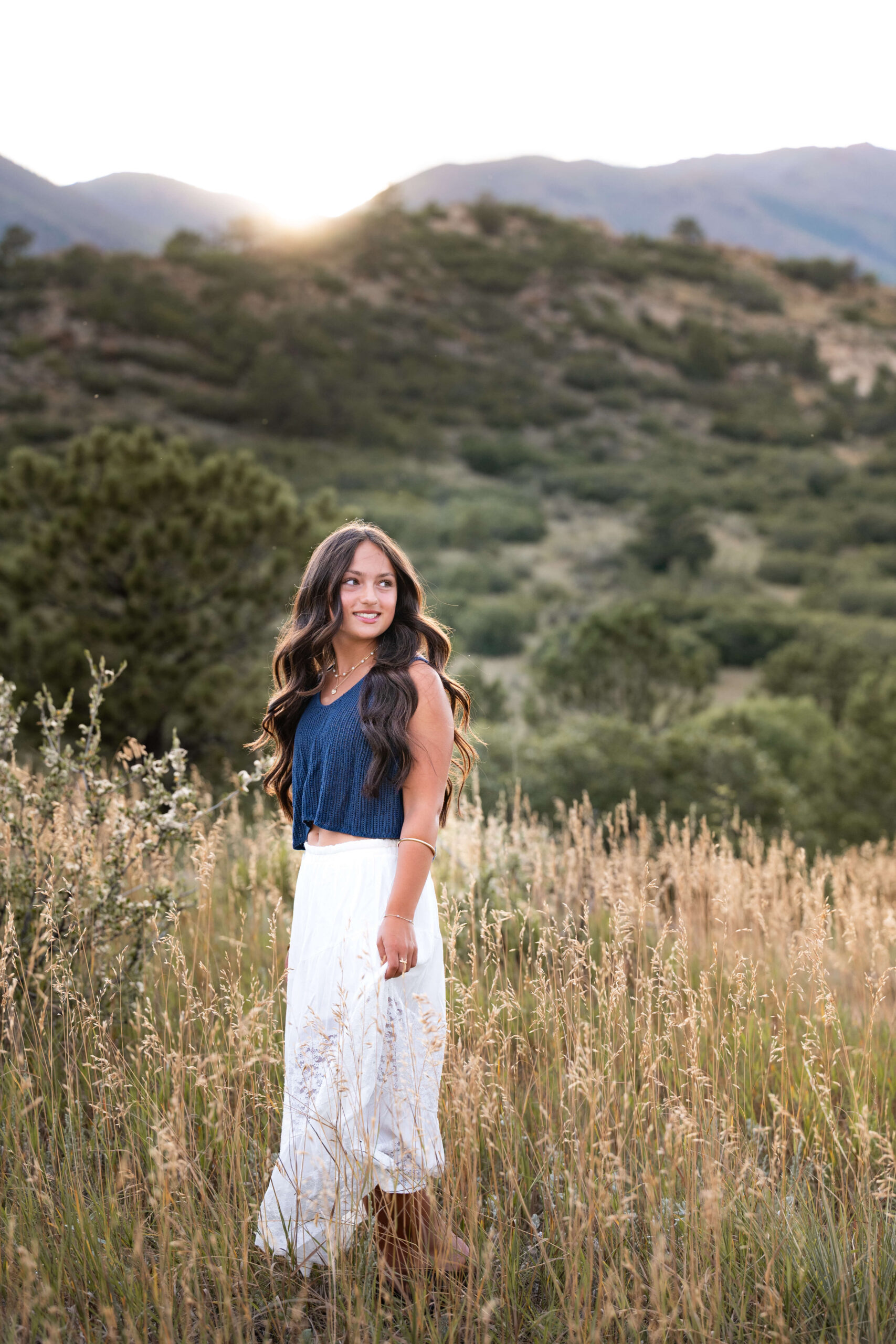 A smiling high school senior in a blue blouse and white skirt walks in tall golden grass on a hillside at sunset after finding nail salons in colorado springs