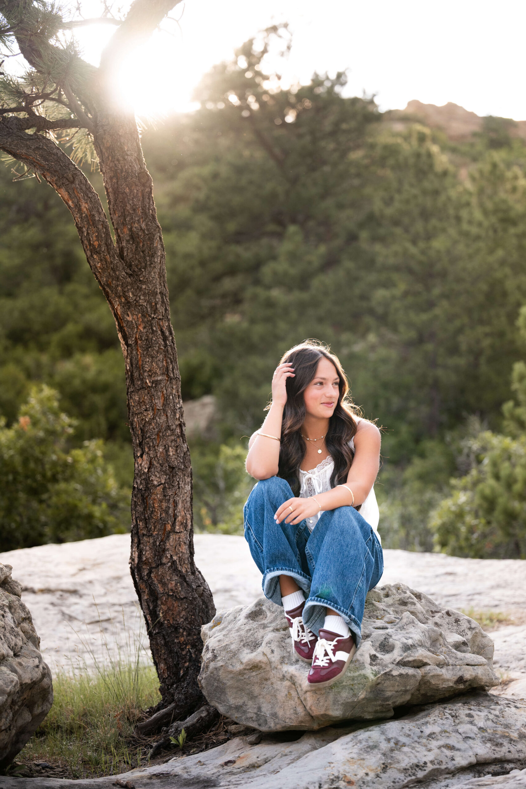 A high school senior in jeans sits on a boulder in the mountains smiling with a hand in her hair after visiting nail salons in colorado springs