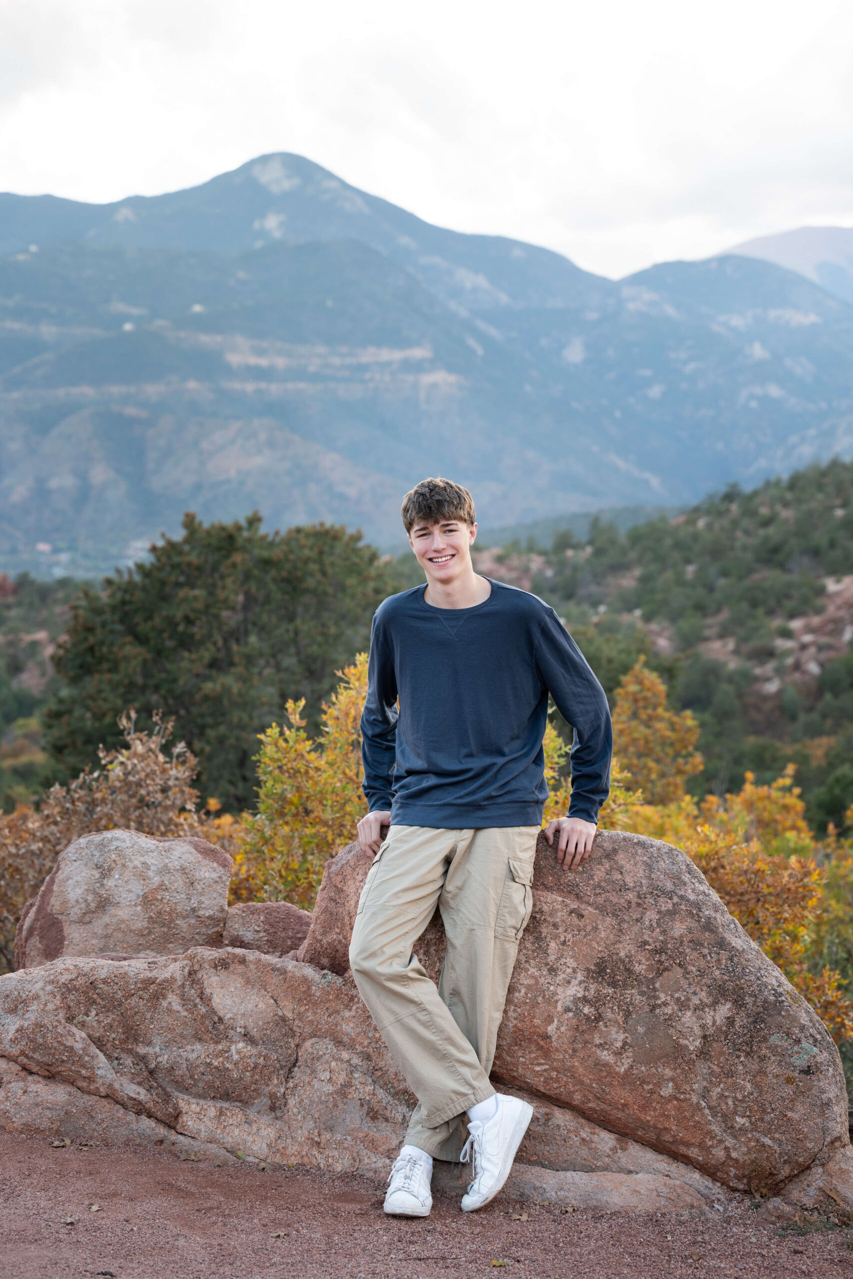 A smiling teenager leans on a boulder in a mountain park at sunset in a blue shirt and khakis