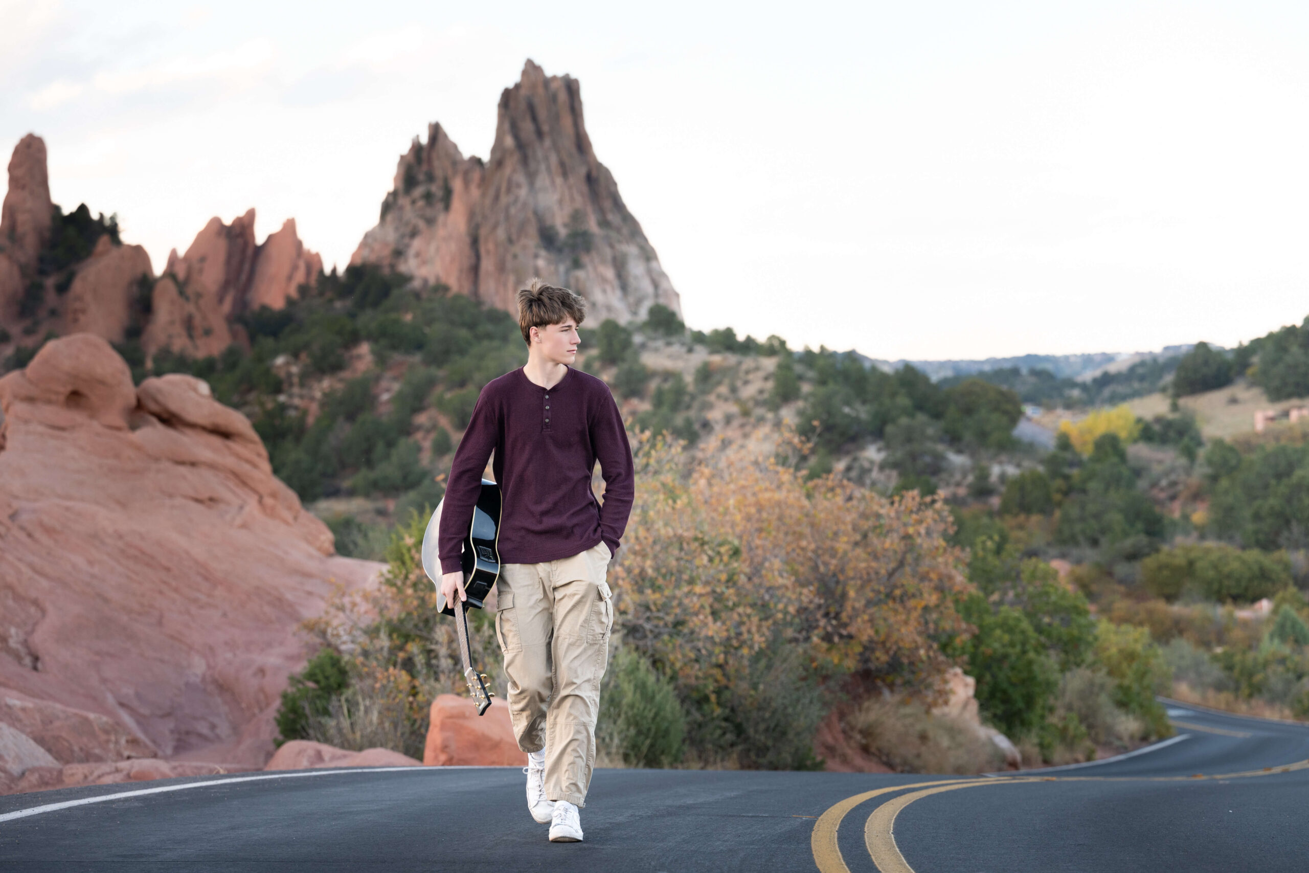 A teenage boy in a maroon shirt walks holding a black guitar on a windy mountain road after finding tutors in colorado springs
