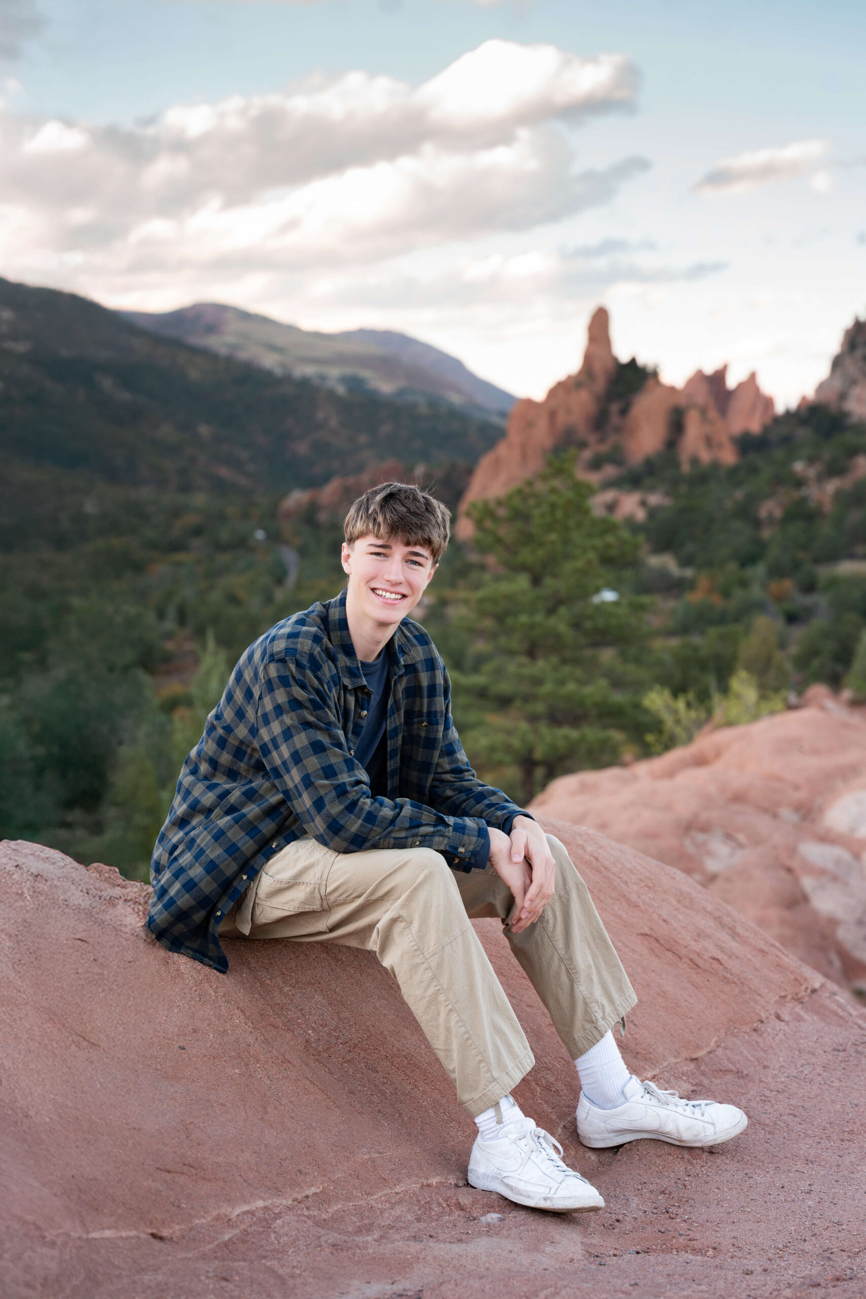 A high school senior sits on red rocks in the mountains in a plaid shirt and khaki pants at sunset after exploring tutors in colorado springs