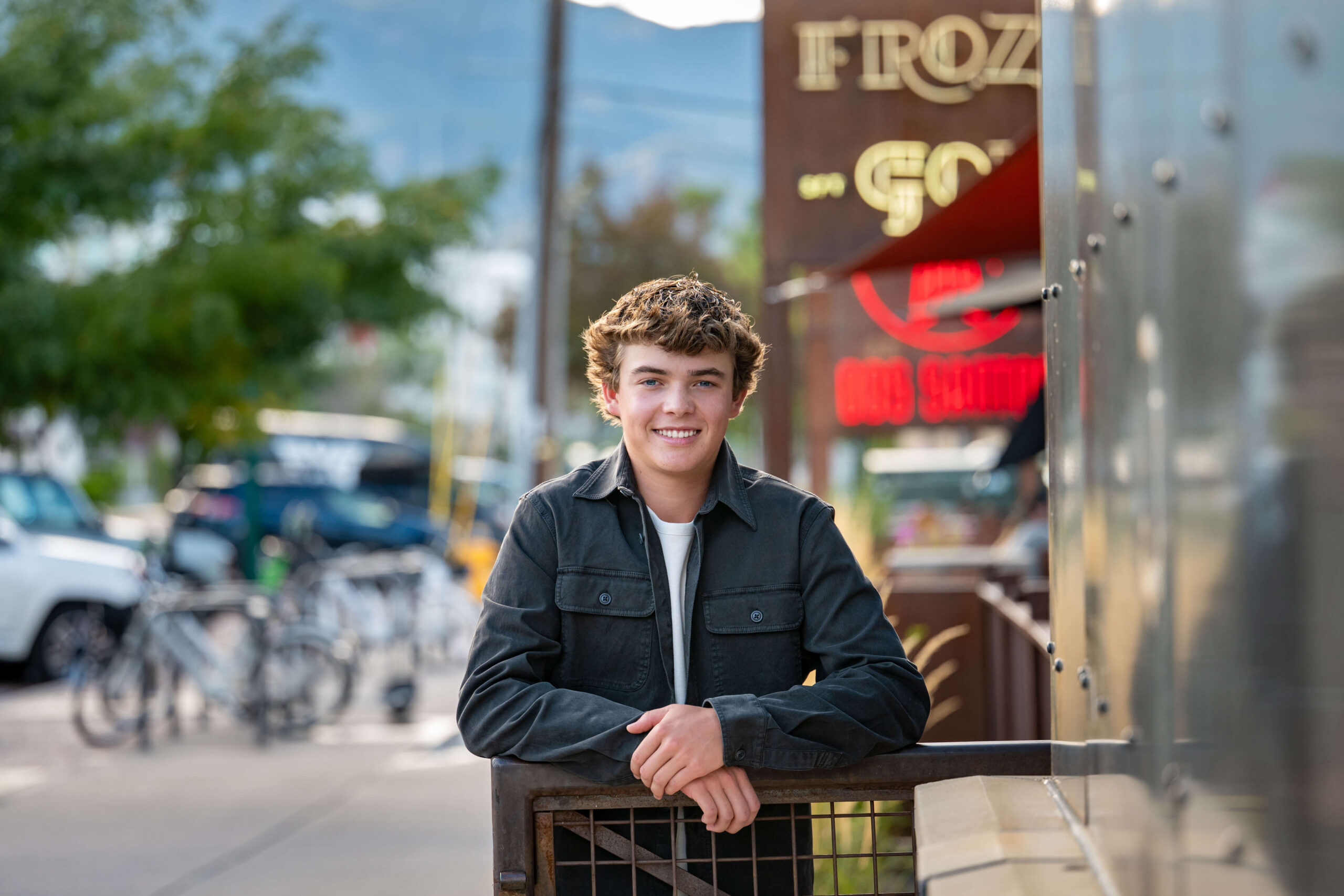 A happy teenage boy in a dark shirt leans on a short metal railing in downtown smiling
