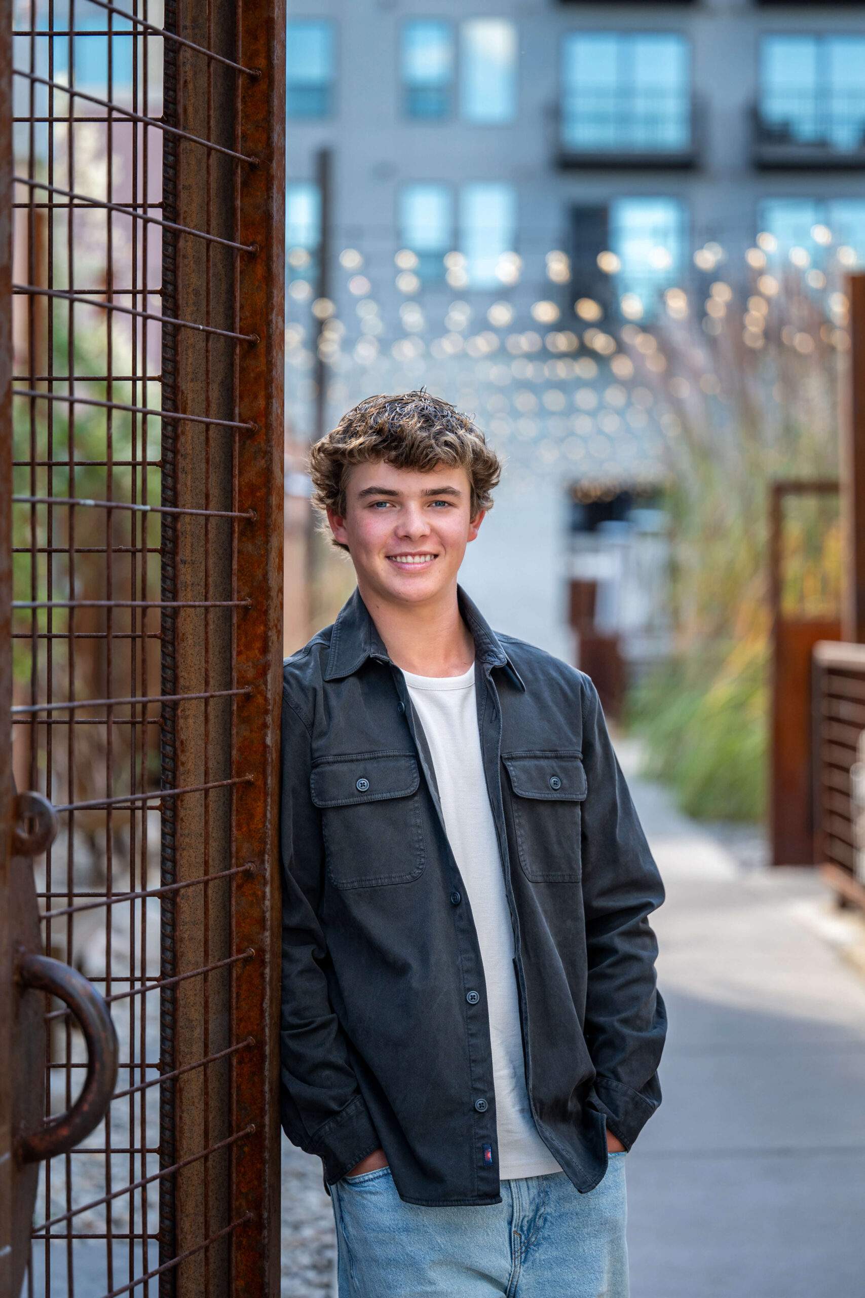 A happy high school senior leans against a rusted metal fence in a patio after exploring tux rental in colorado springs