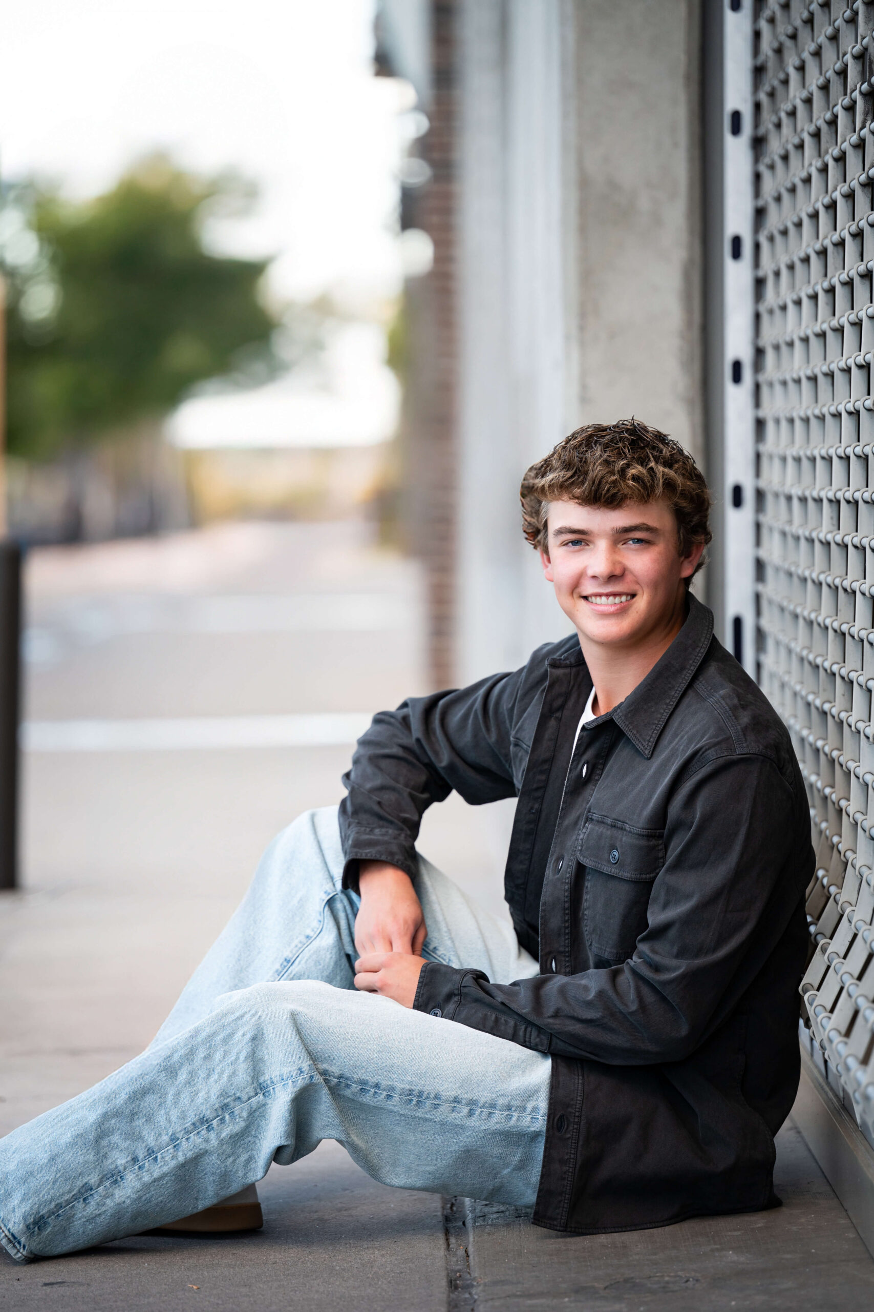 A high school senior in a black shirt and jeans sits against a roll down gate smiling after finding tux rental in colorado springs