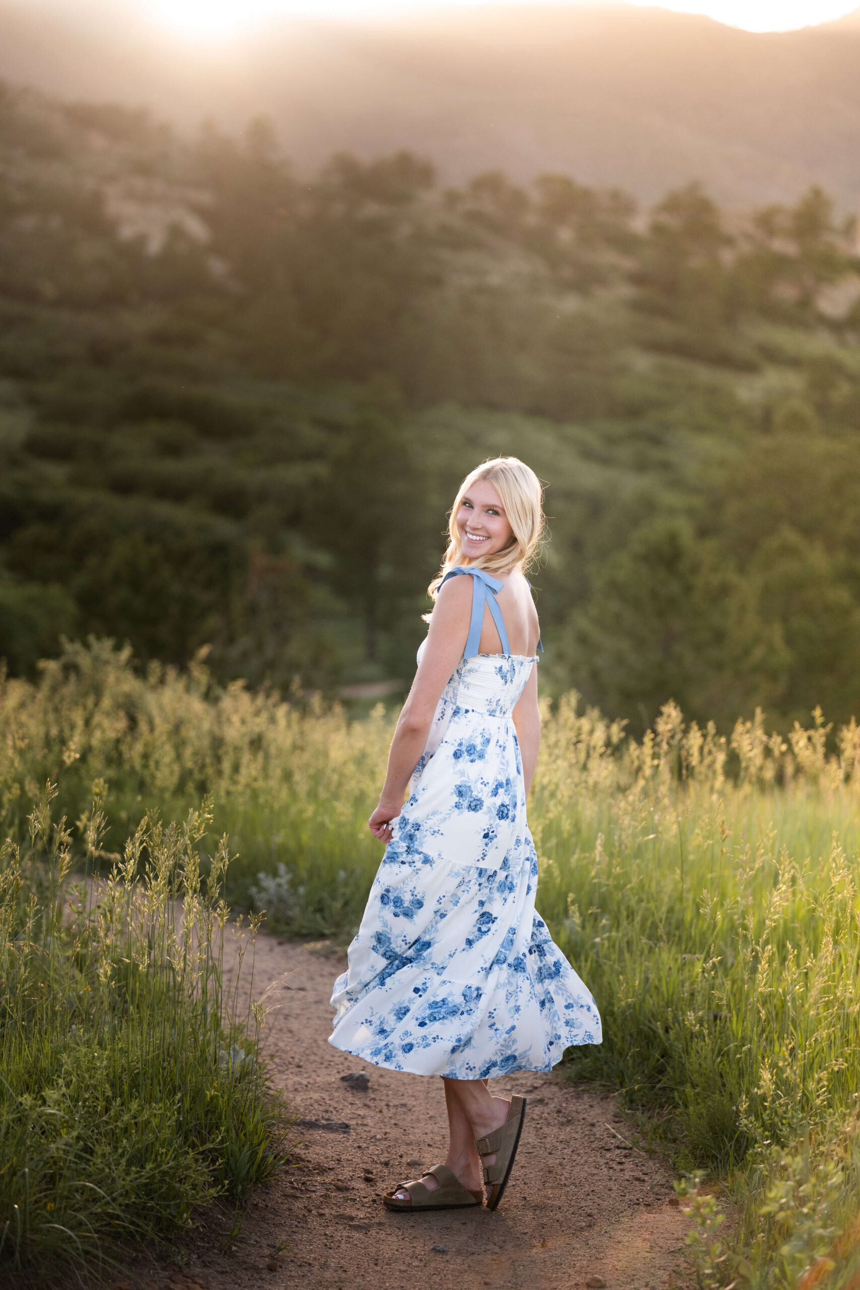 A happy blonde woman in a blue floral print dress smiles over her shoulder in a park trail at sunset