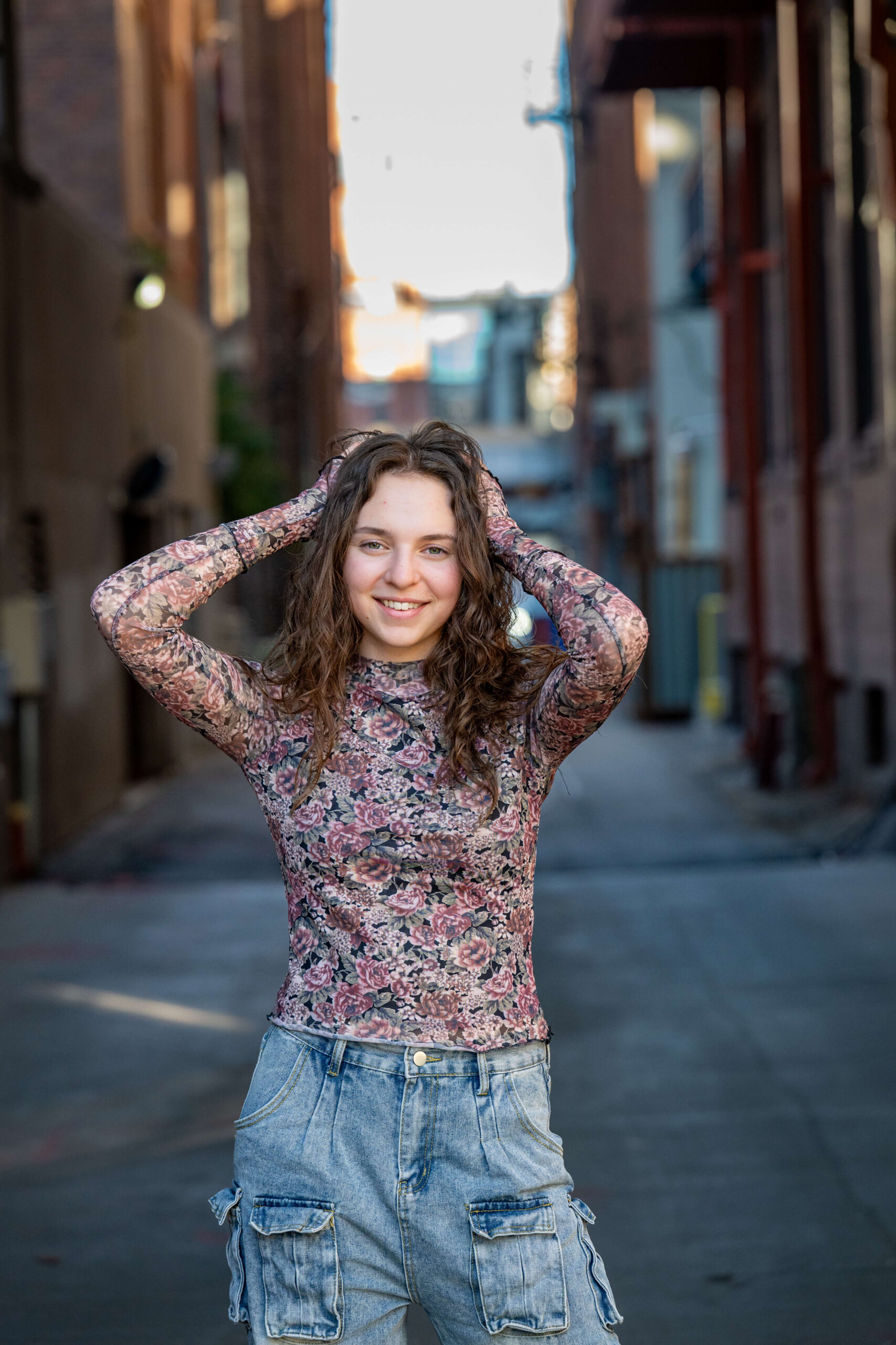 A teenager in a flower print blouse stands in an alley smiling with hands in her hair after finding Colorado Springs Boutiques