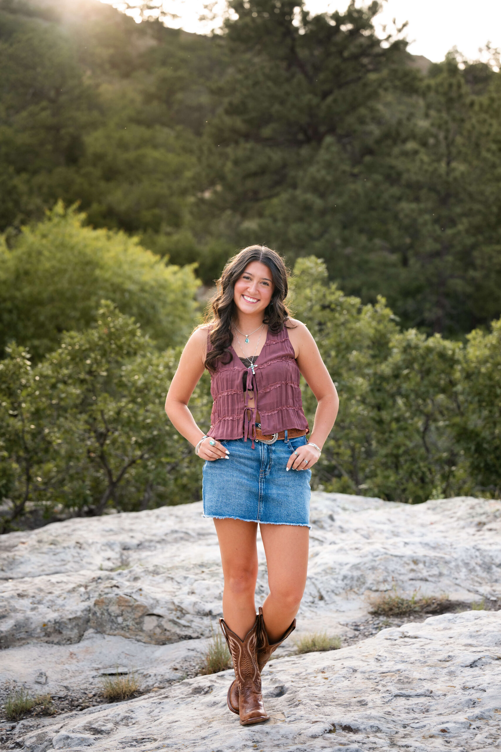 A smiling high school senior stands on a mountain with trees behind her, in a jean skirt and cowboy boots.