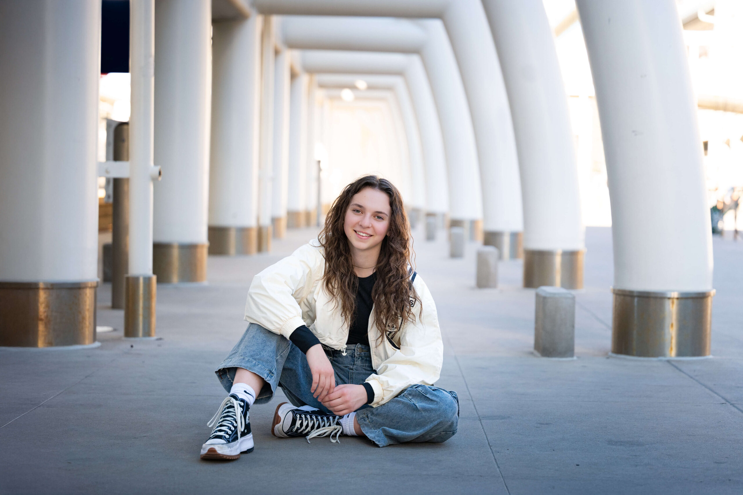 A high school senior sits in a row of columns, smiling in jeans and a white jacket at the college fair of colorado springs.