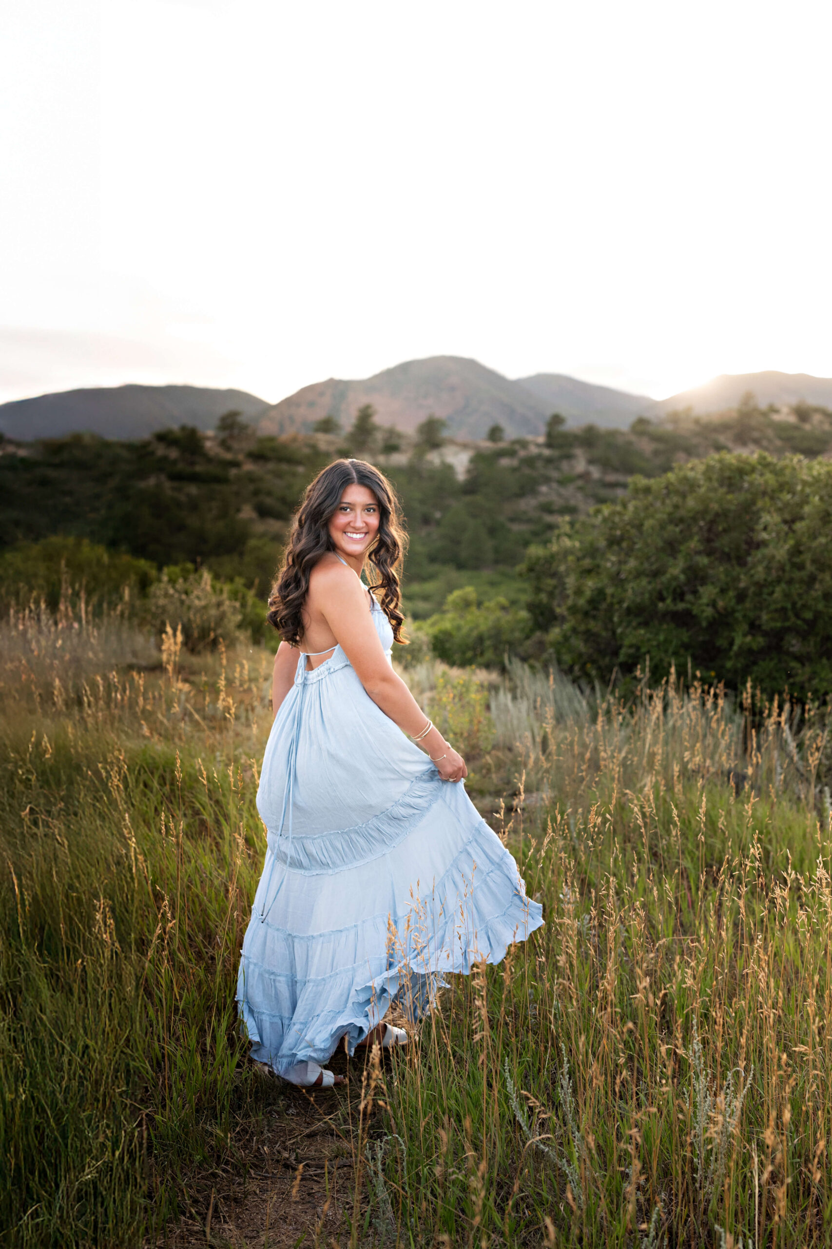 A happy high school senior in a long blue dress walks through a field of tall golden grass in the mountains for colorado springs senior photos.