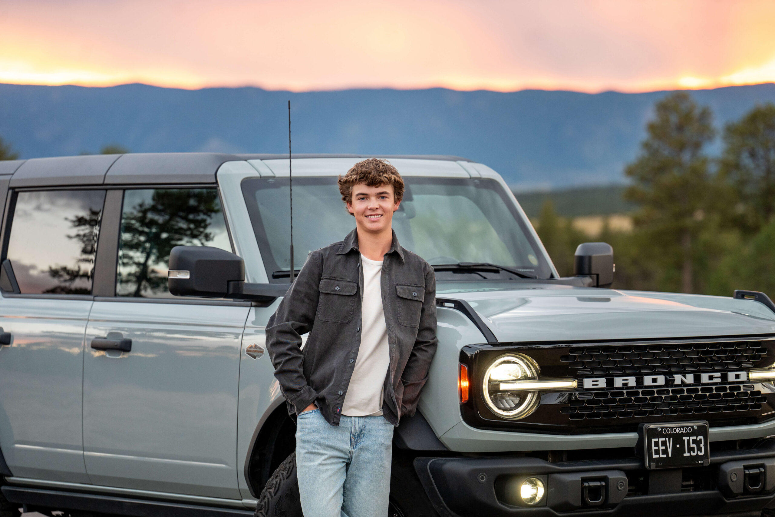 A high school senior leans against his Ford Bronco at sunset in the mountains.