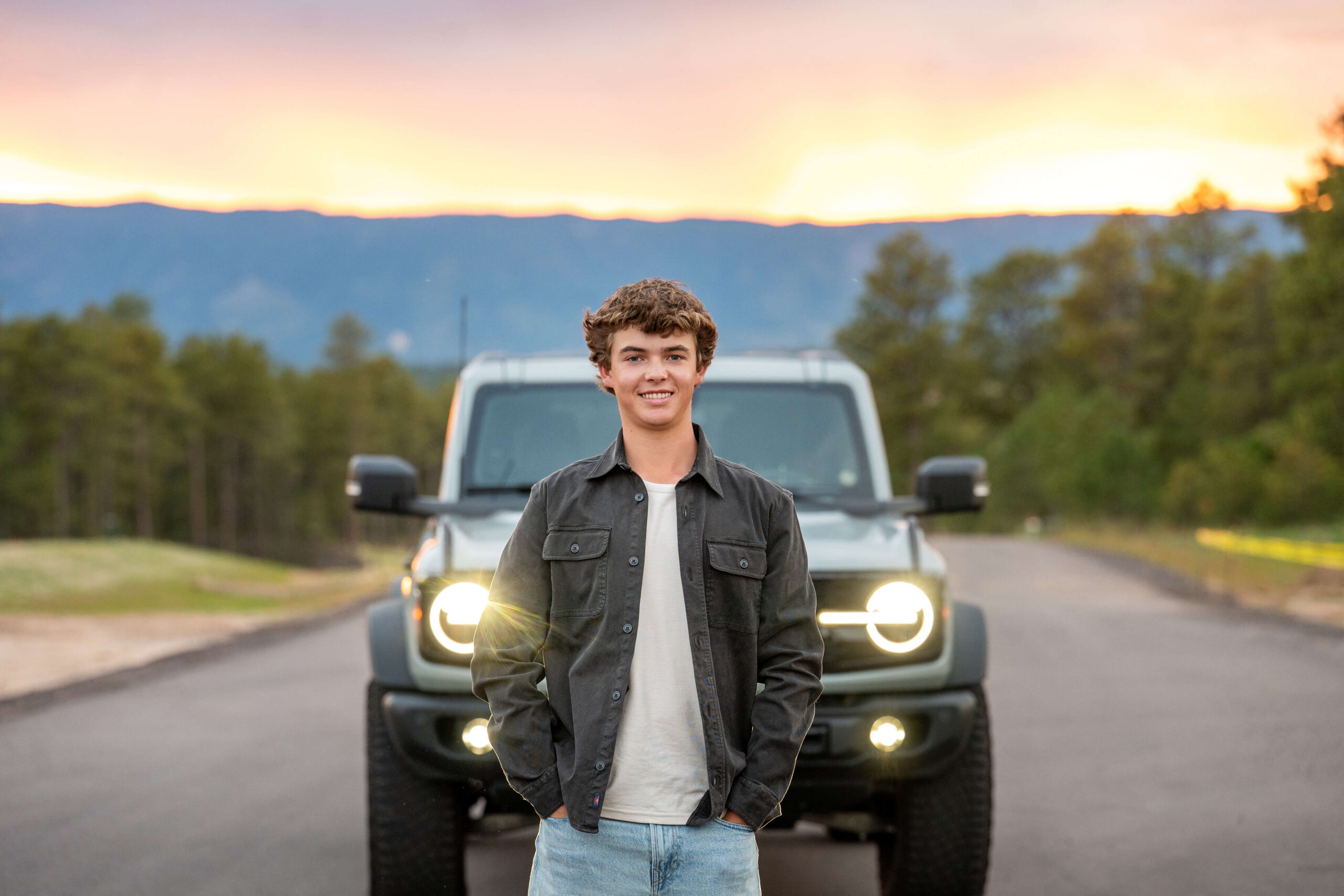 A high school senior stands in front of his car parked on a mountain road at sunset after looking for graduation party venues in colorado springs.